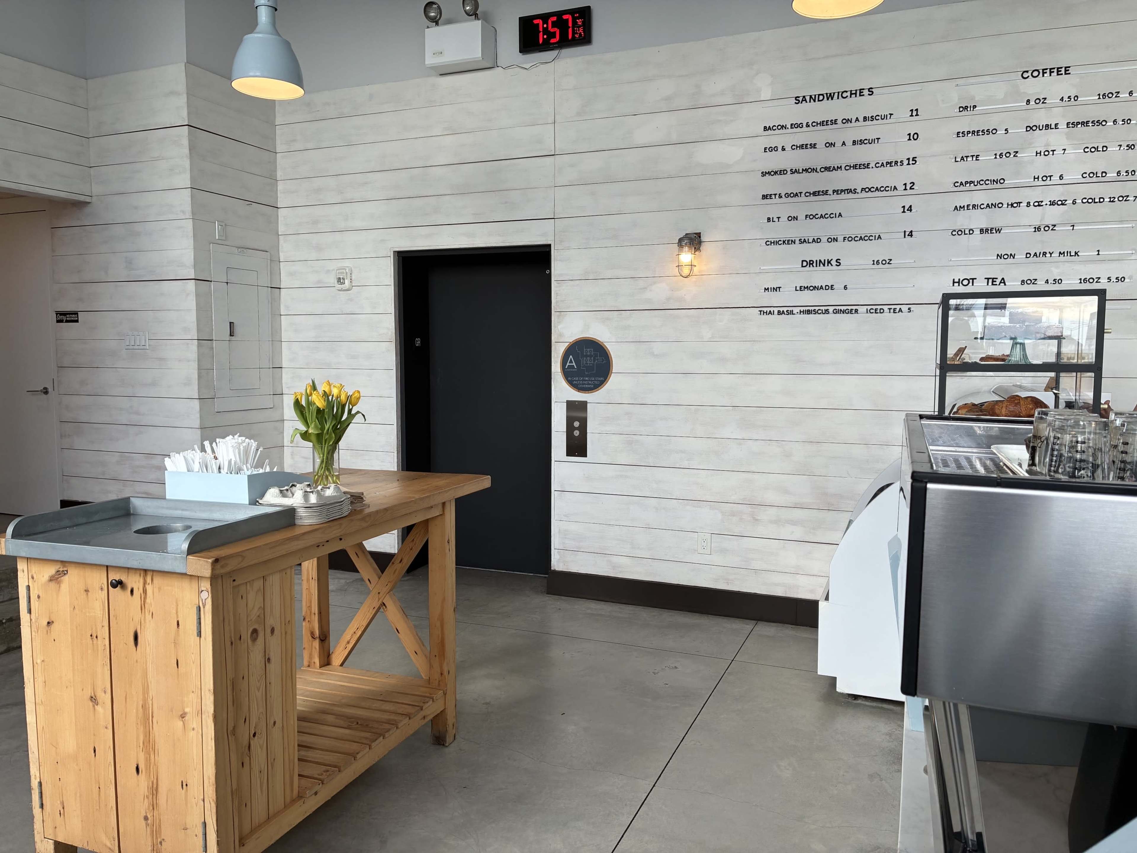 The image shows a modern café interior with a wooden service cart, a coffee machine, and an elevator door, along with a chalkboard menu on the wall.