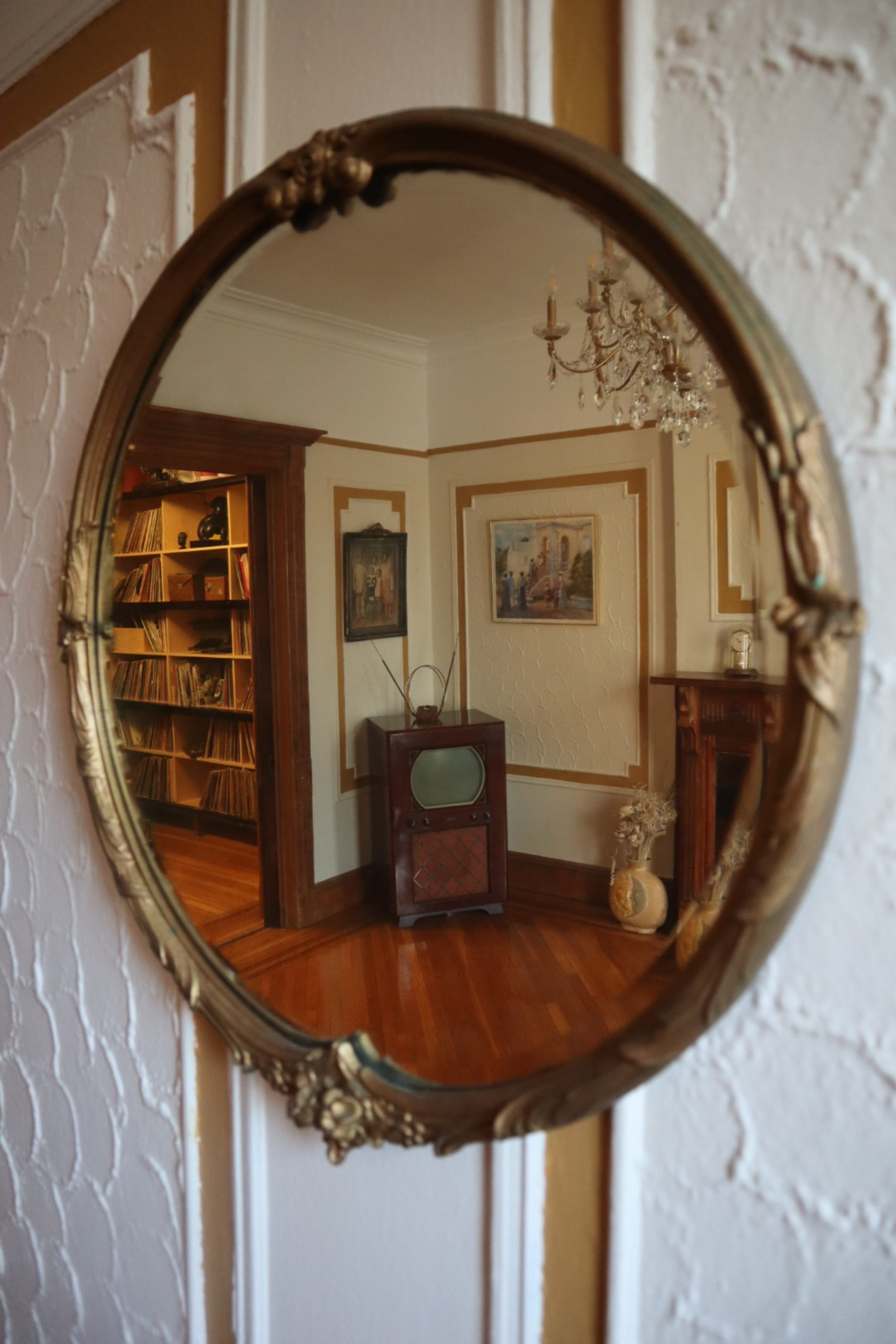 A round, ornate mirror reflects a room with a vintage television, bookshelf, and framed paintings.