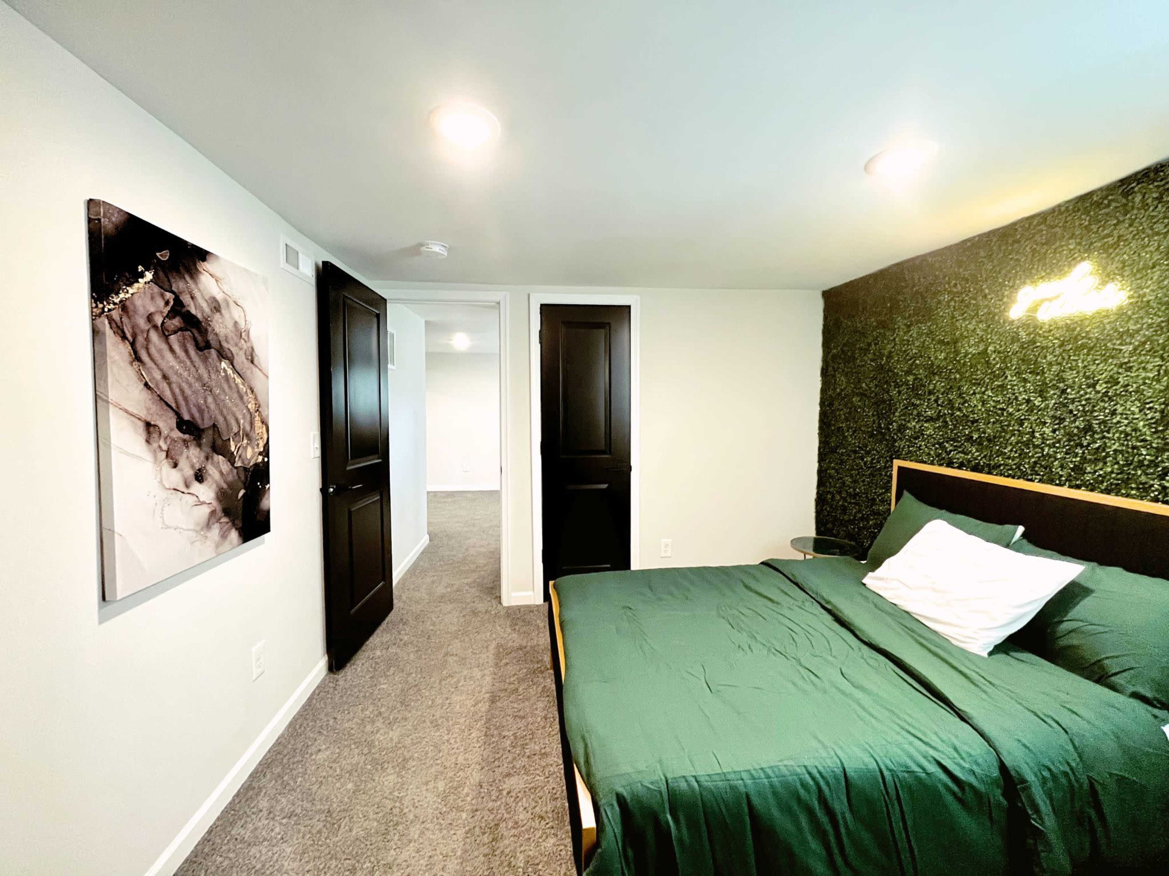 A modern bedroom featuring a green bedspread, a wall with artificial greenery, and a doorway leading to another room.