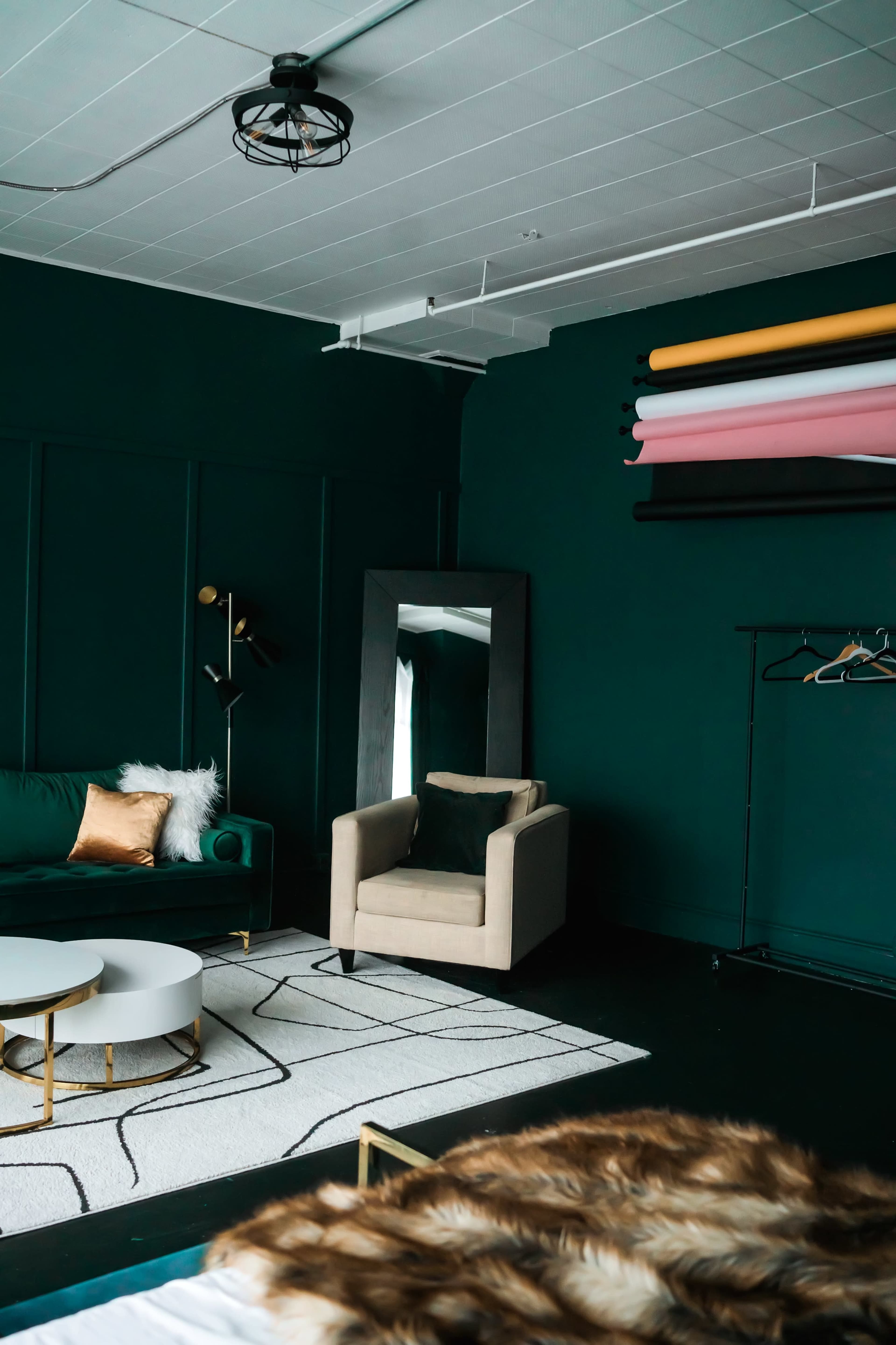 The image shows a stylish room featuring a dark green wall, a green sofa with decorative pillows, a beige armchair, a large mirror, a round coffee table, and rolls of colored paper mounted on the wall.