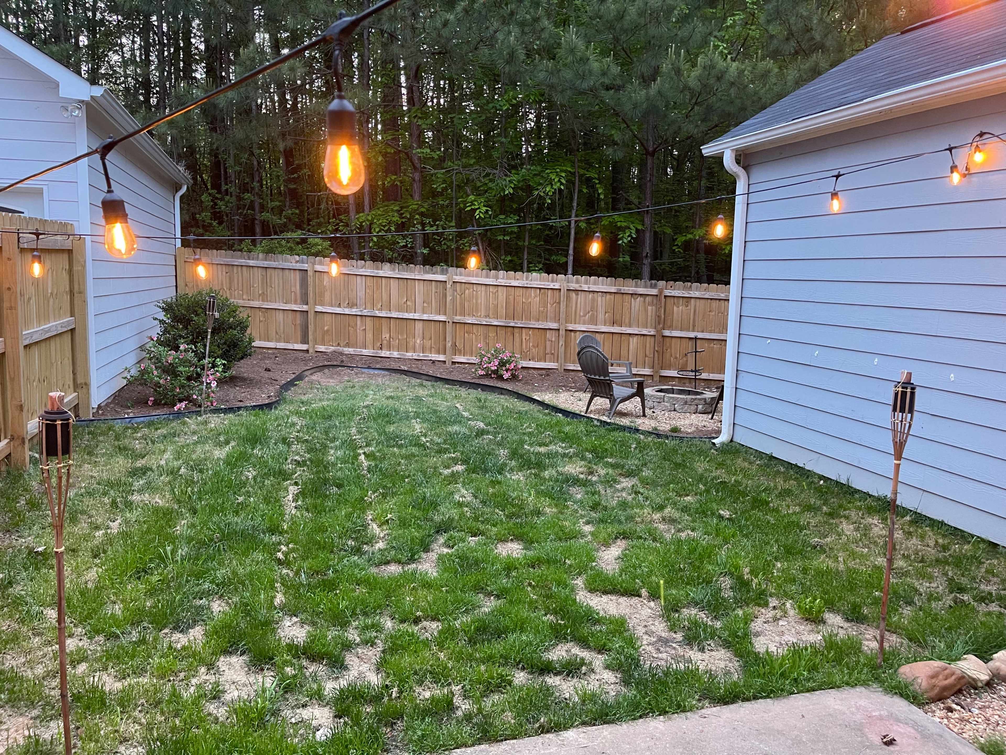 The image shows a backyard with a grassy area lit by string lights and bordered by wooden fencing, featuring a seating area and landscaping with flower beds.