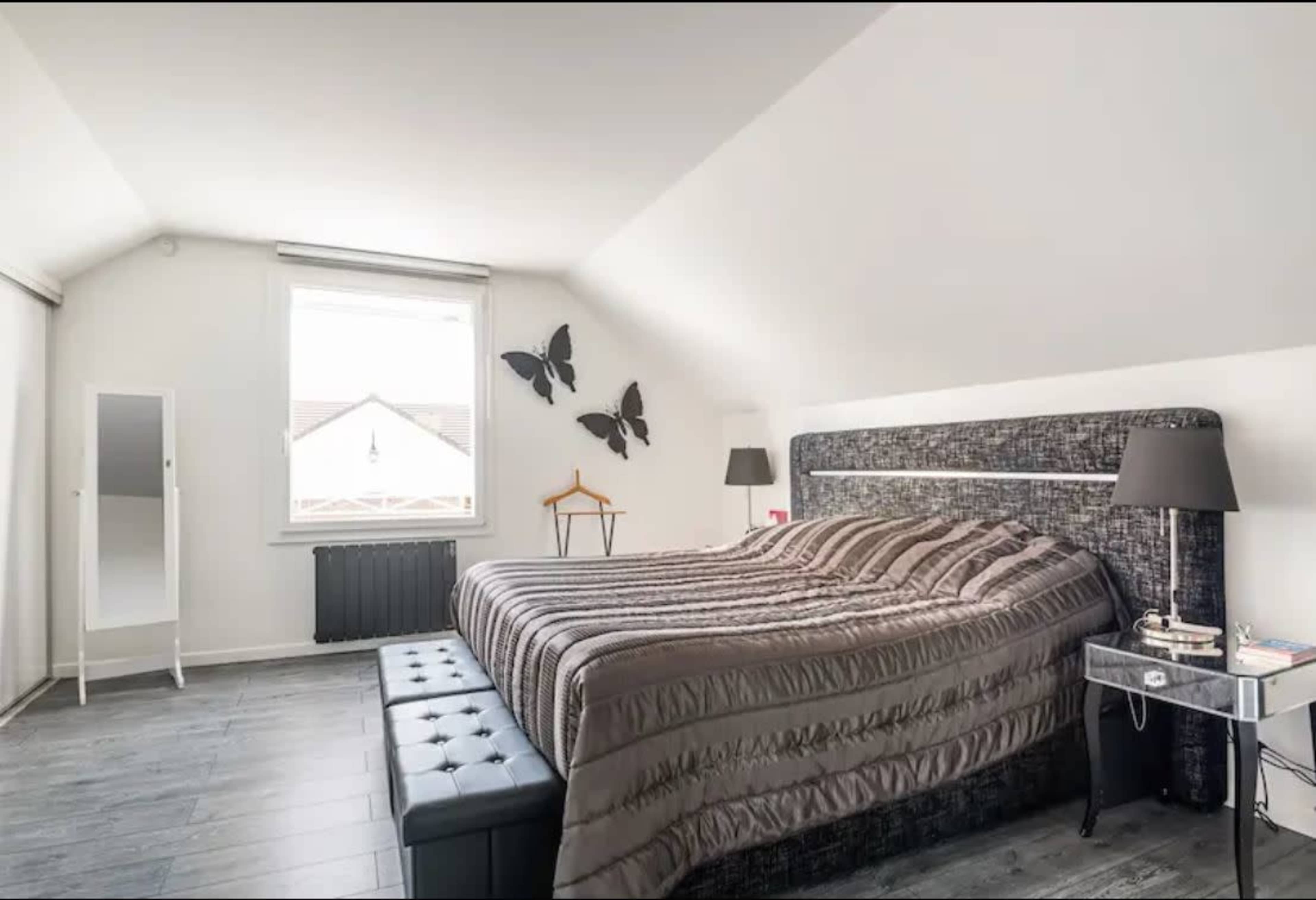 The image shows a modern bedroom featuring a large bed with patterned bedding, a black storage bench at the foot of the bed, a full-length mirror, and decorative butterflies on the wall.