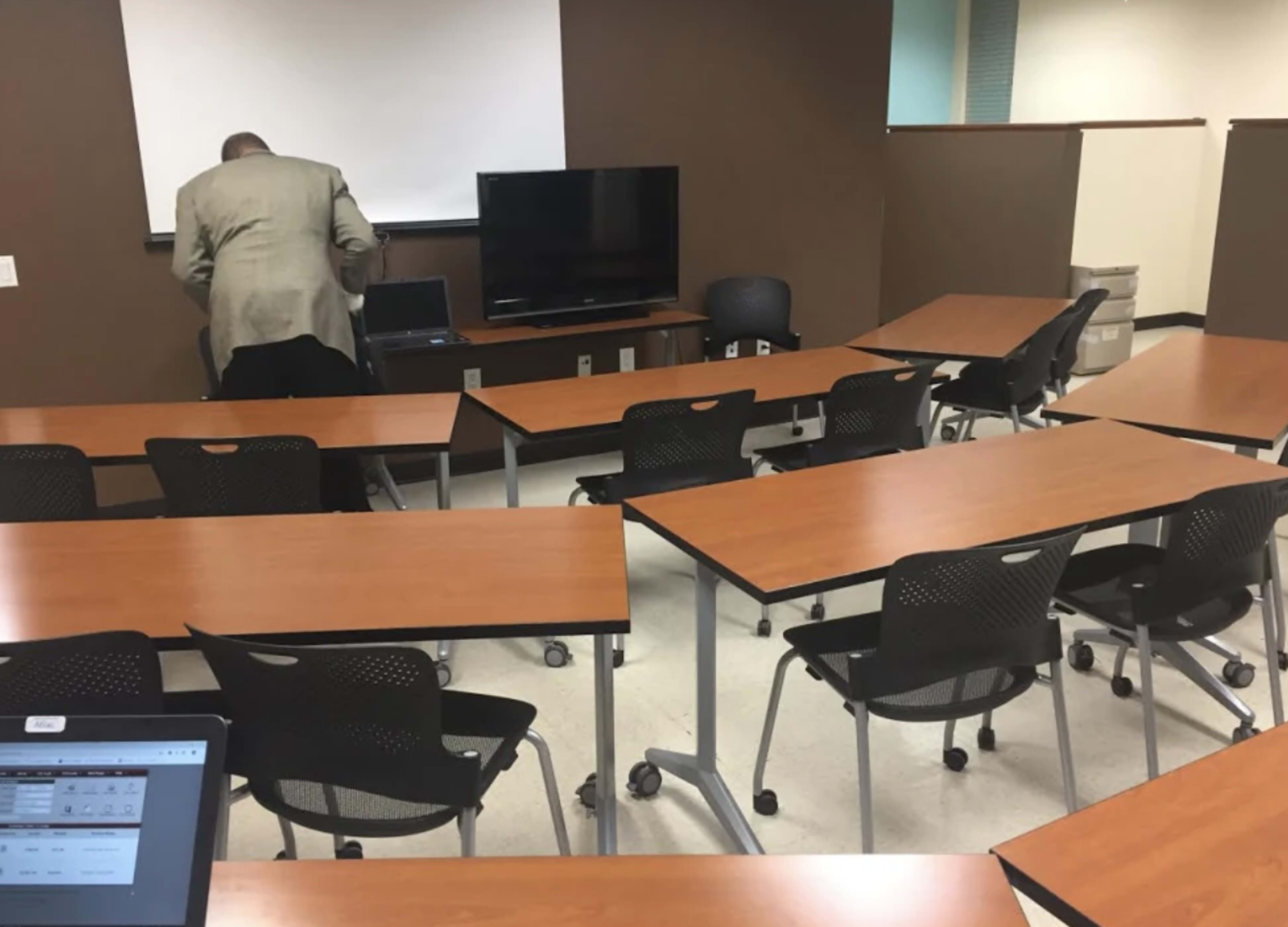 A man in a suit stands near a laptop in an empty classroom with movable tables and chairs arranged in a U-shape.