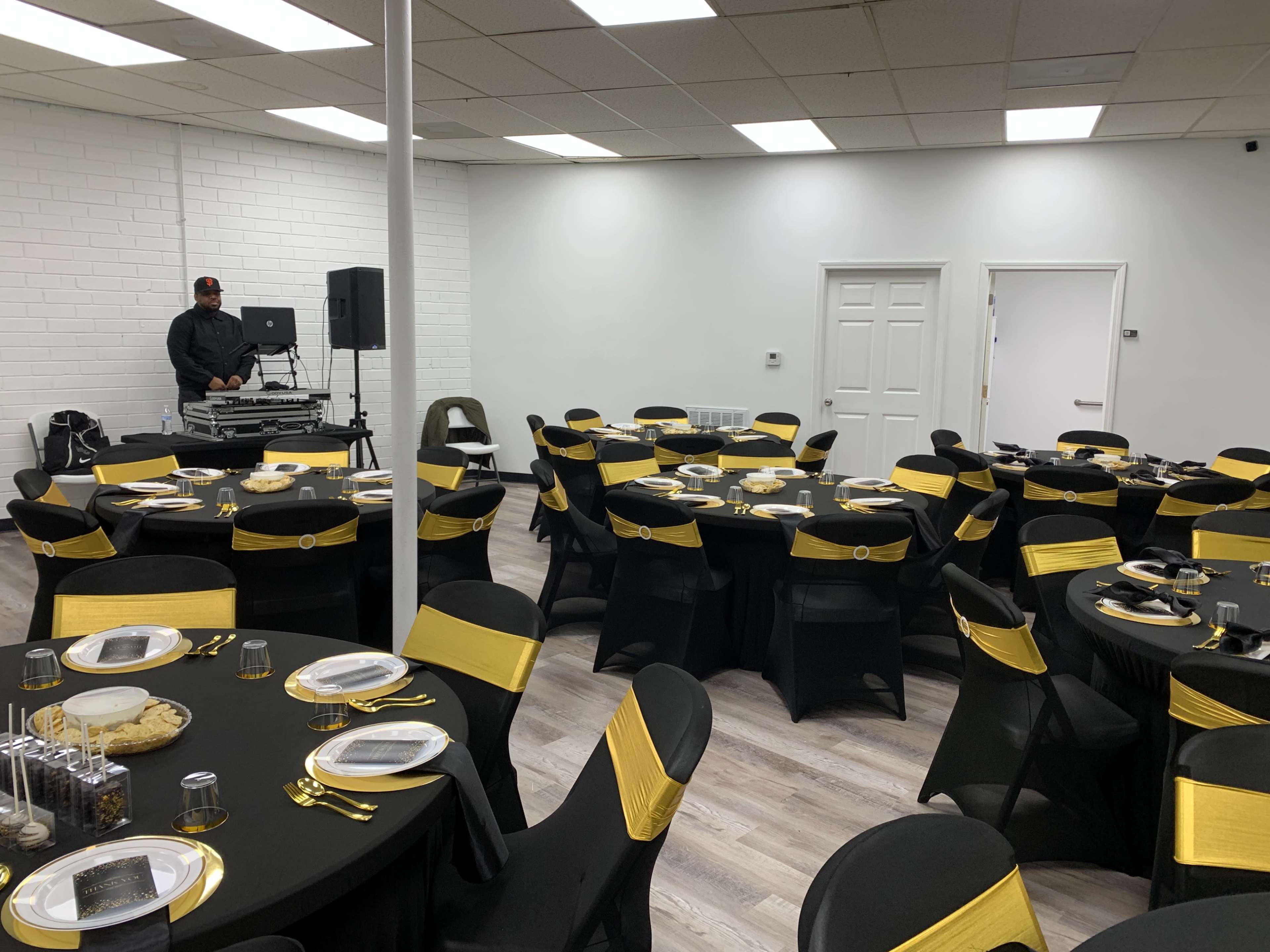 A large event space features black tables with yellow accents, each set with plates and utensils, while a DJ stands in the corner.