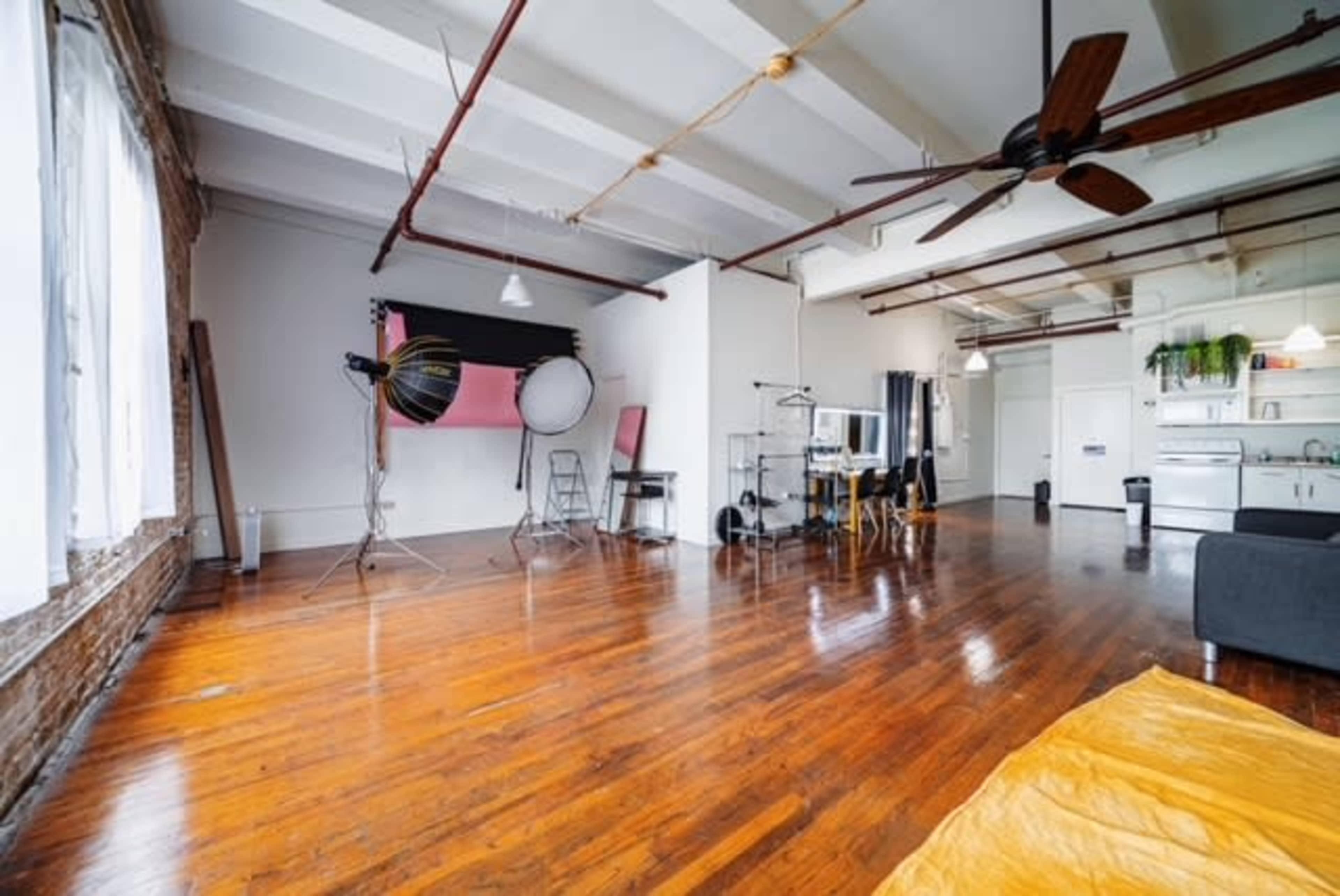 A spacious, well-lit studio with wooden floors, featuring photography equipment, a small kitchen area, and minimal furnishings.