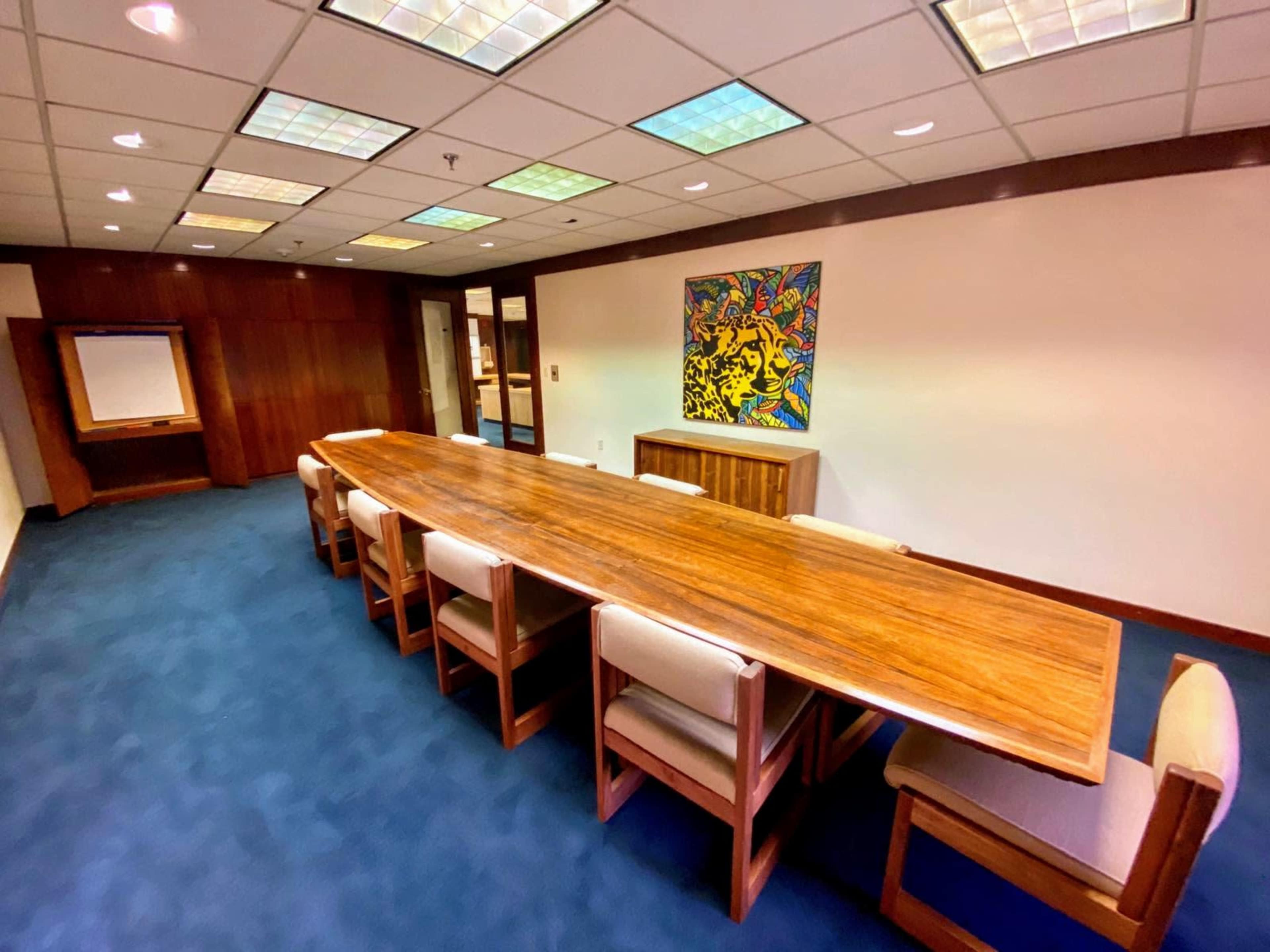 A large wooden conference table with eight chairs surrounds it in a well-lit meeting room featuring colorful artwork on the wall.