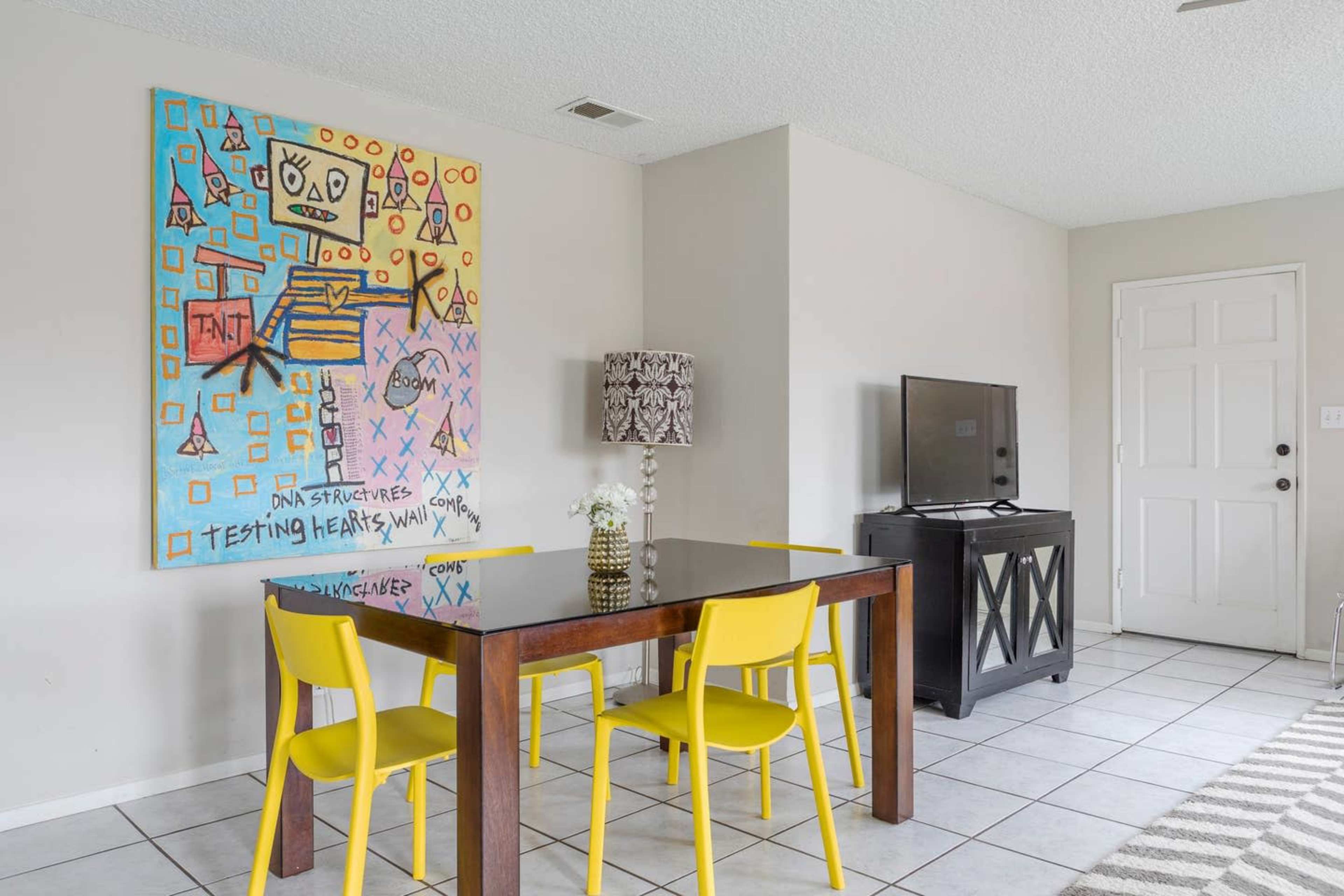 A dining area features a black table with two yellow chairs, a colorful abstract painting on the wall, and a television on a cabinet beside a white door.
