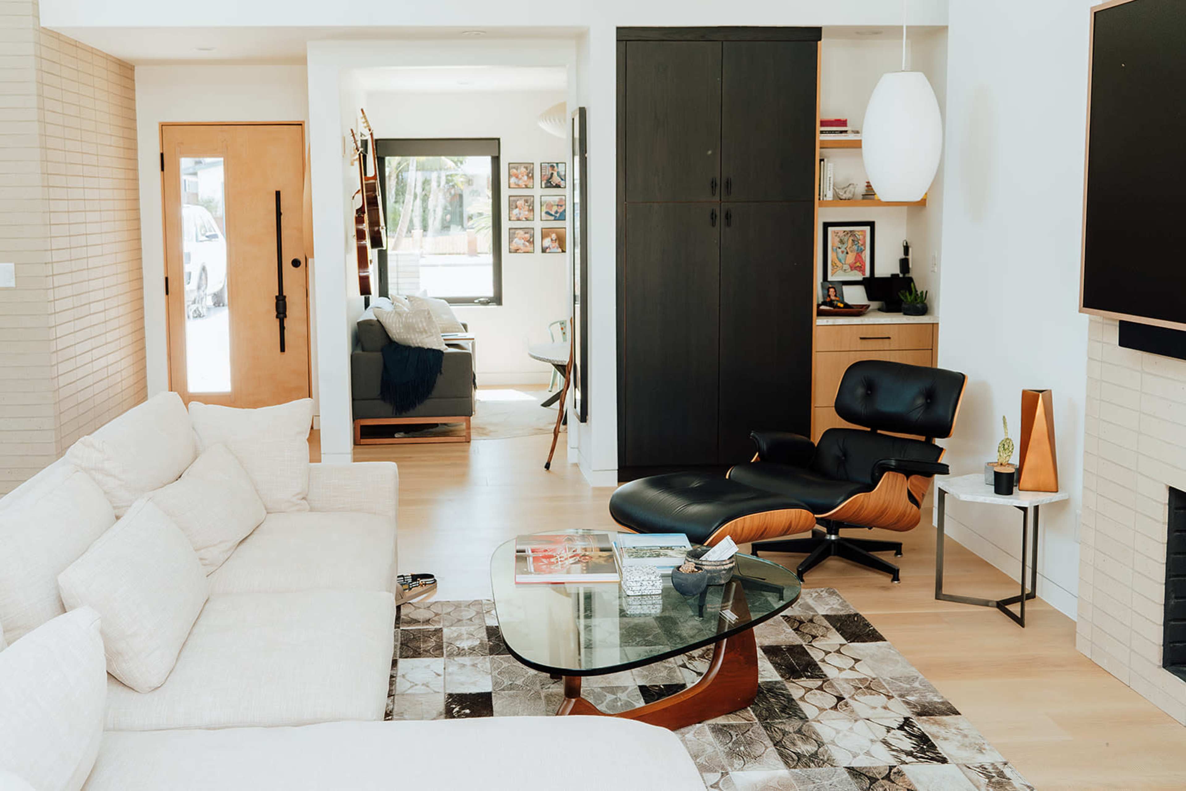 The image shows a modern living room with a white sectional sofa, a glass coffee table, and a black lounge chair, complemented by walls adorned with framed artwork and a light-colored interior palette.