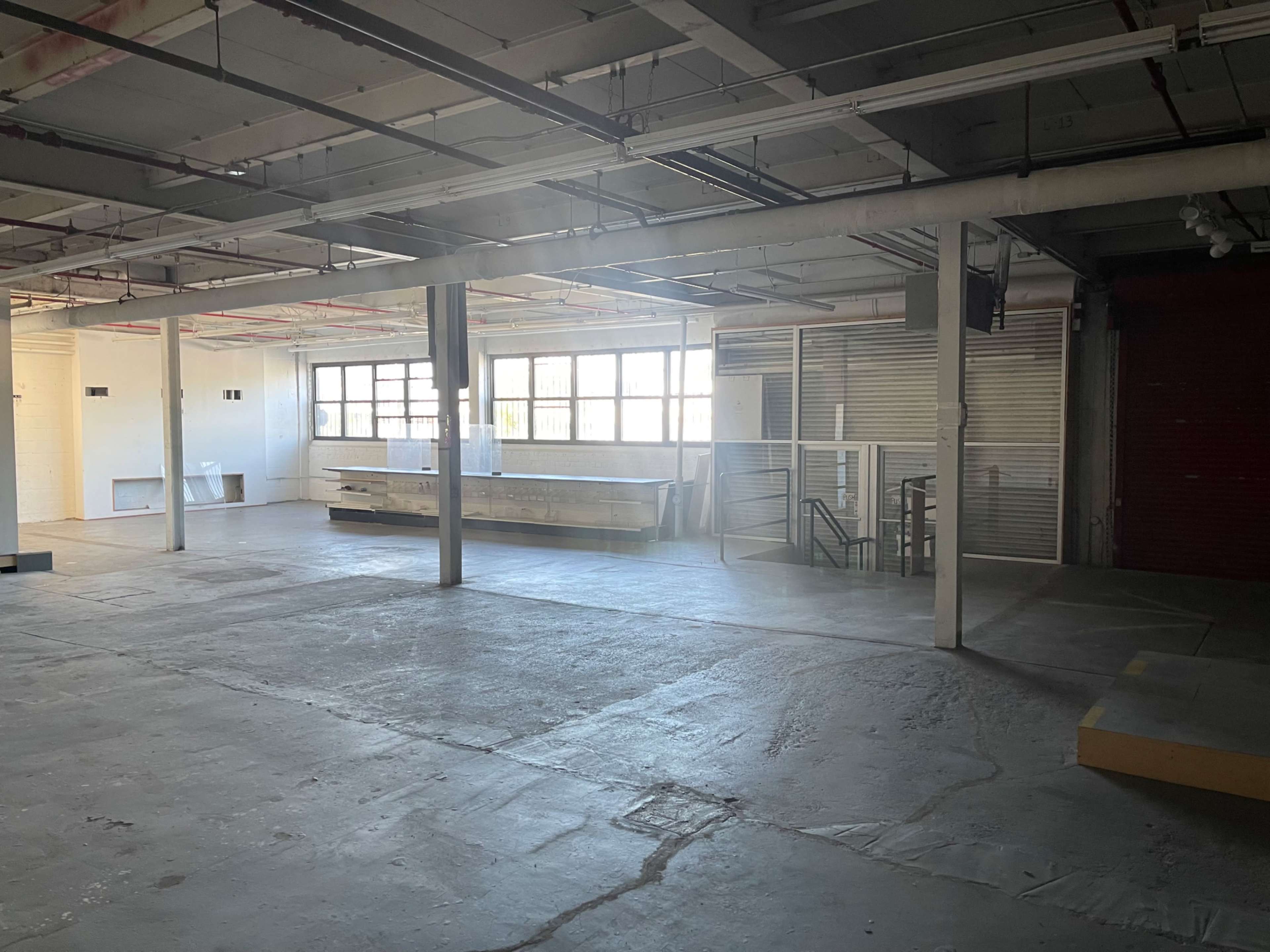 The image shows an empty commercial space with concrete floors, large windows, and a staircase in the background.