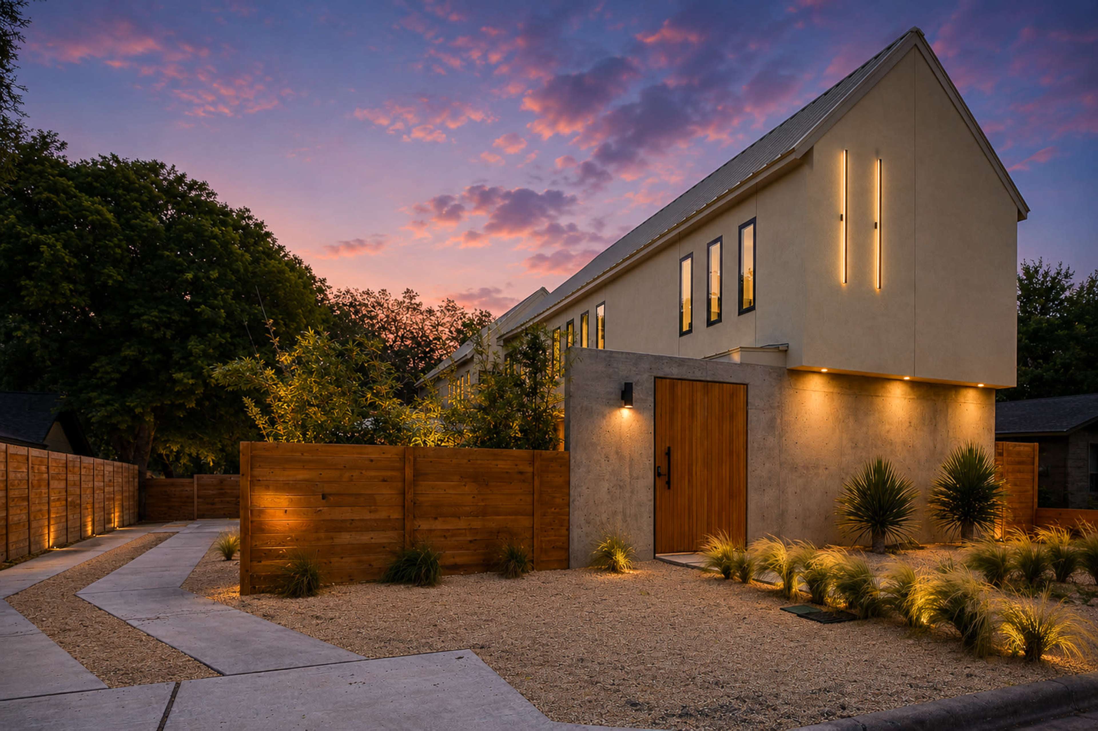 A modern house with a concrete exterior and wooden accents is illuminated at dusk, situated near a gravel pathway and landscaped with low shrubs.