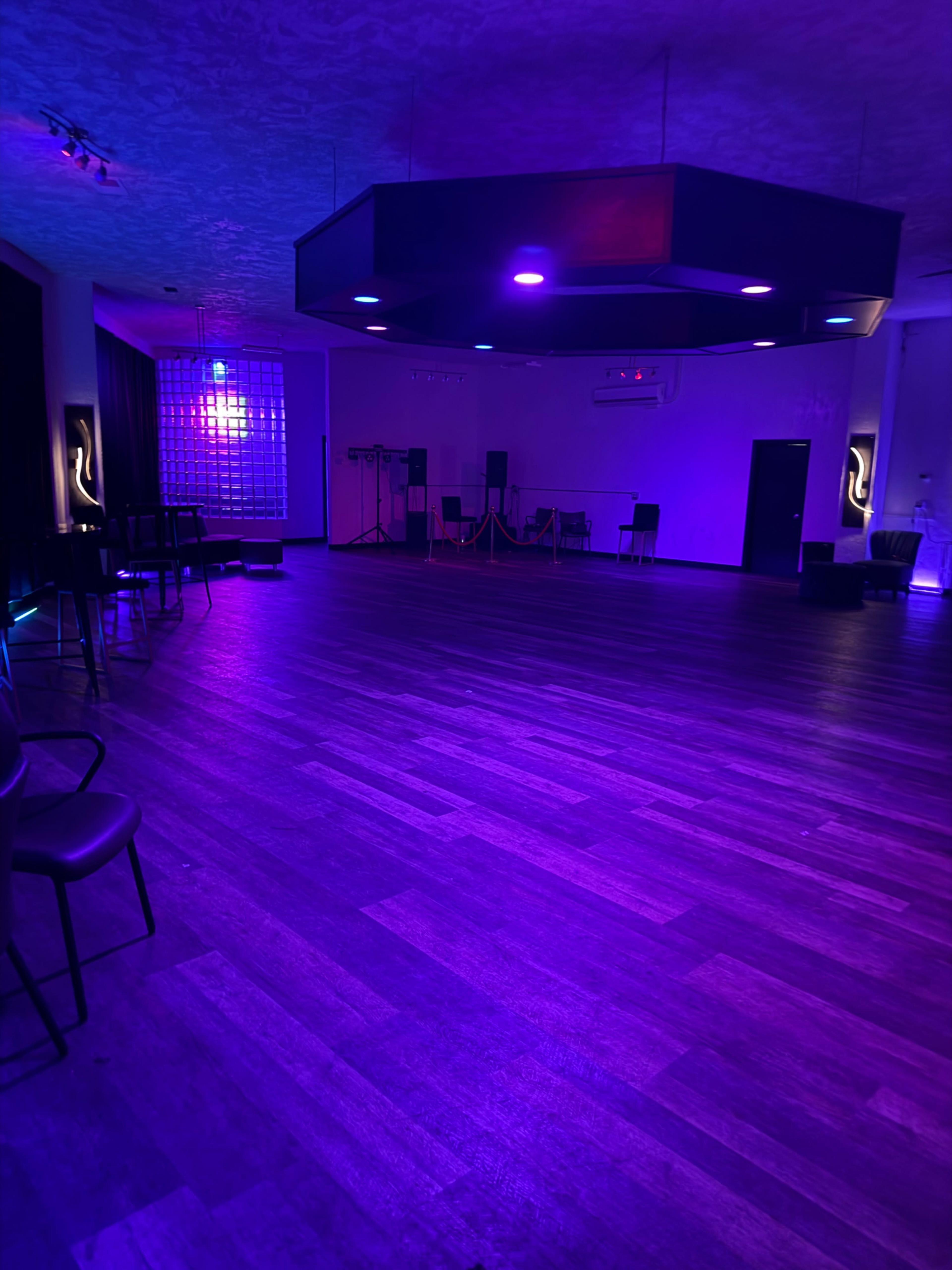The image shows an empty, dimly lit event space with purple lighting, wooden flooring, and a bar area along one side.
