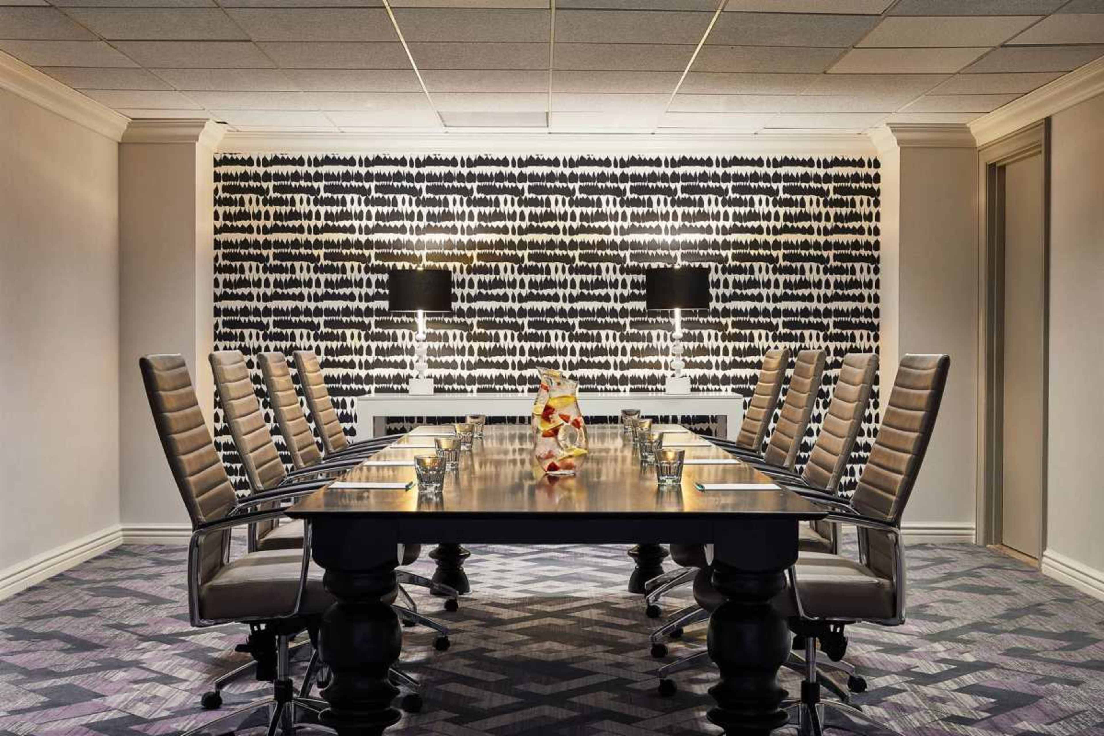 A large conference room features a long dark wooden table surrounded by ergonomic chairs, with a patterned wall illuminated by two lamps.