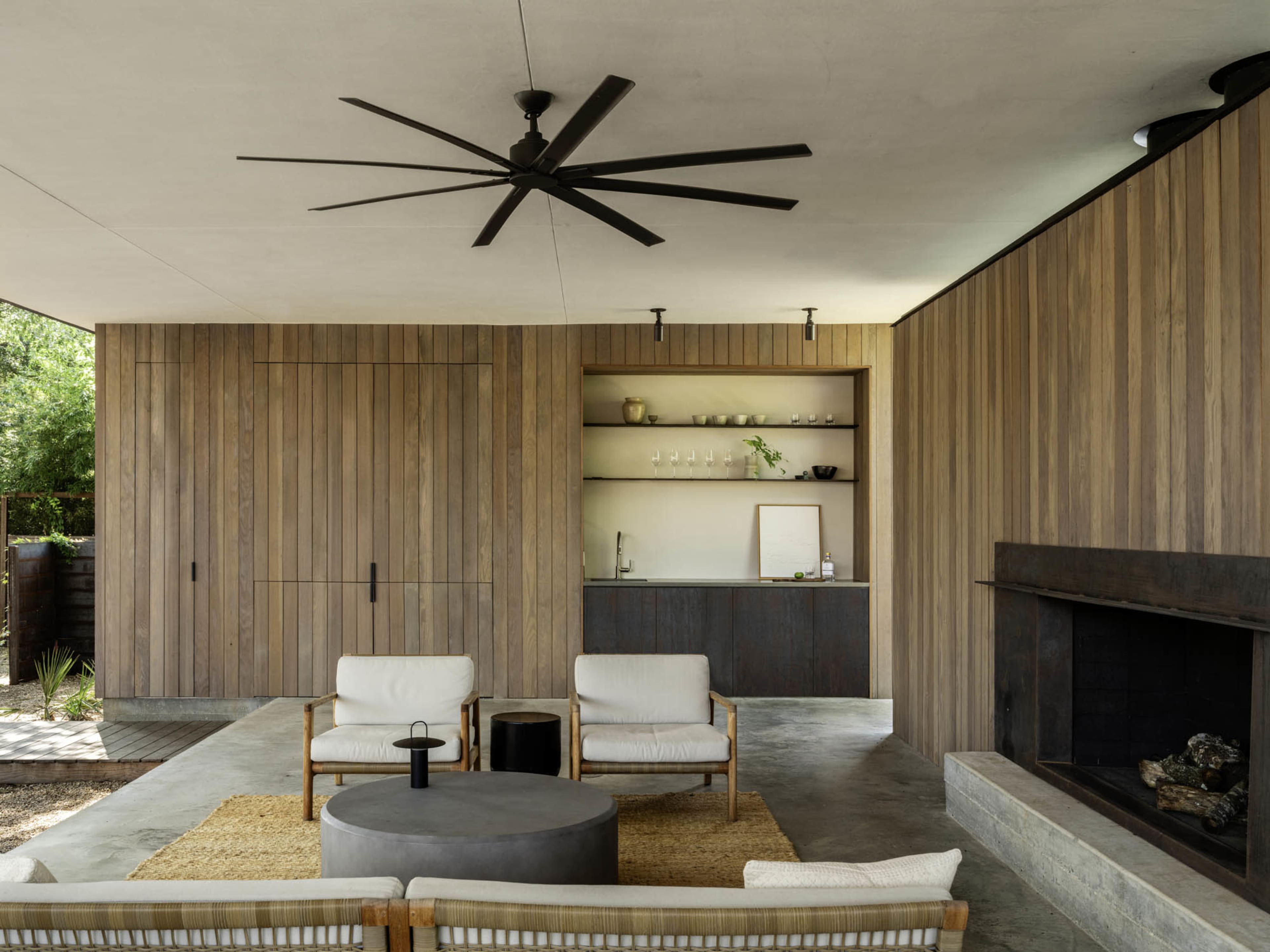 The image shows a modern outdoor living space featuring two white armchairs, a round coffee table, and a sleek kitchen area with wooden paneling and a fireplace.