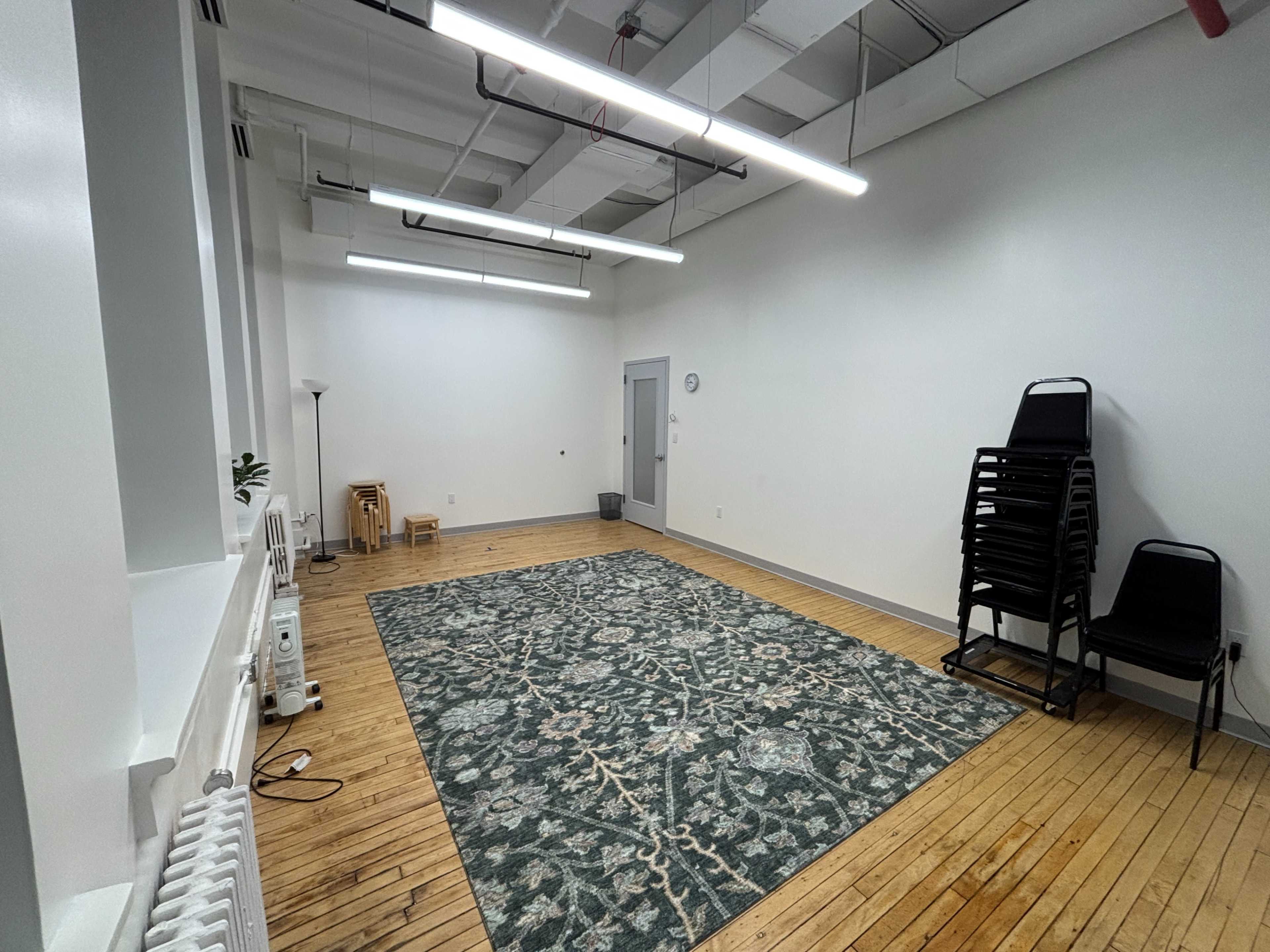 The image shows a sparsely furnished room with a large area rug on the wooden floor, three stacked chairs in one corner, and a small lamp against the wall.