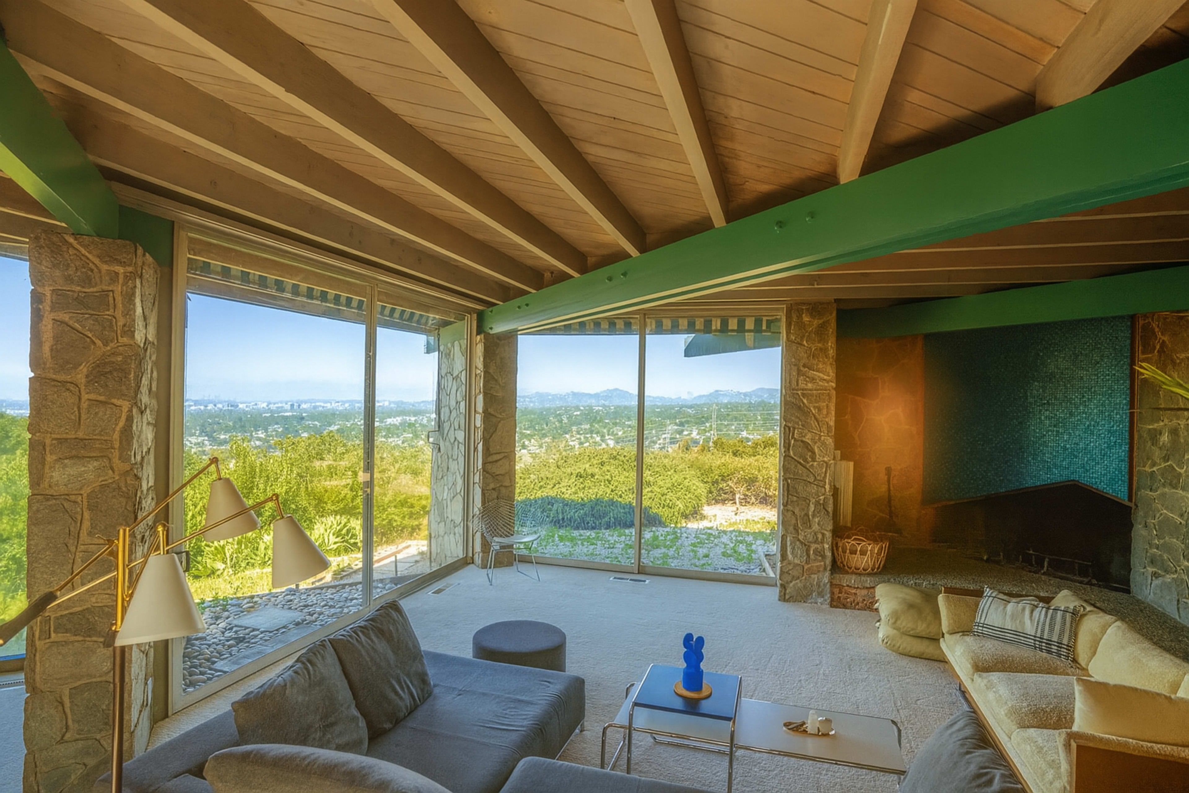 The image shows a modern living room with large windows overlooking a landscape, featuring a stone fireplace and minimalist furniture.