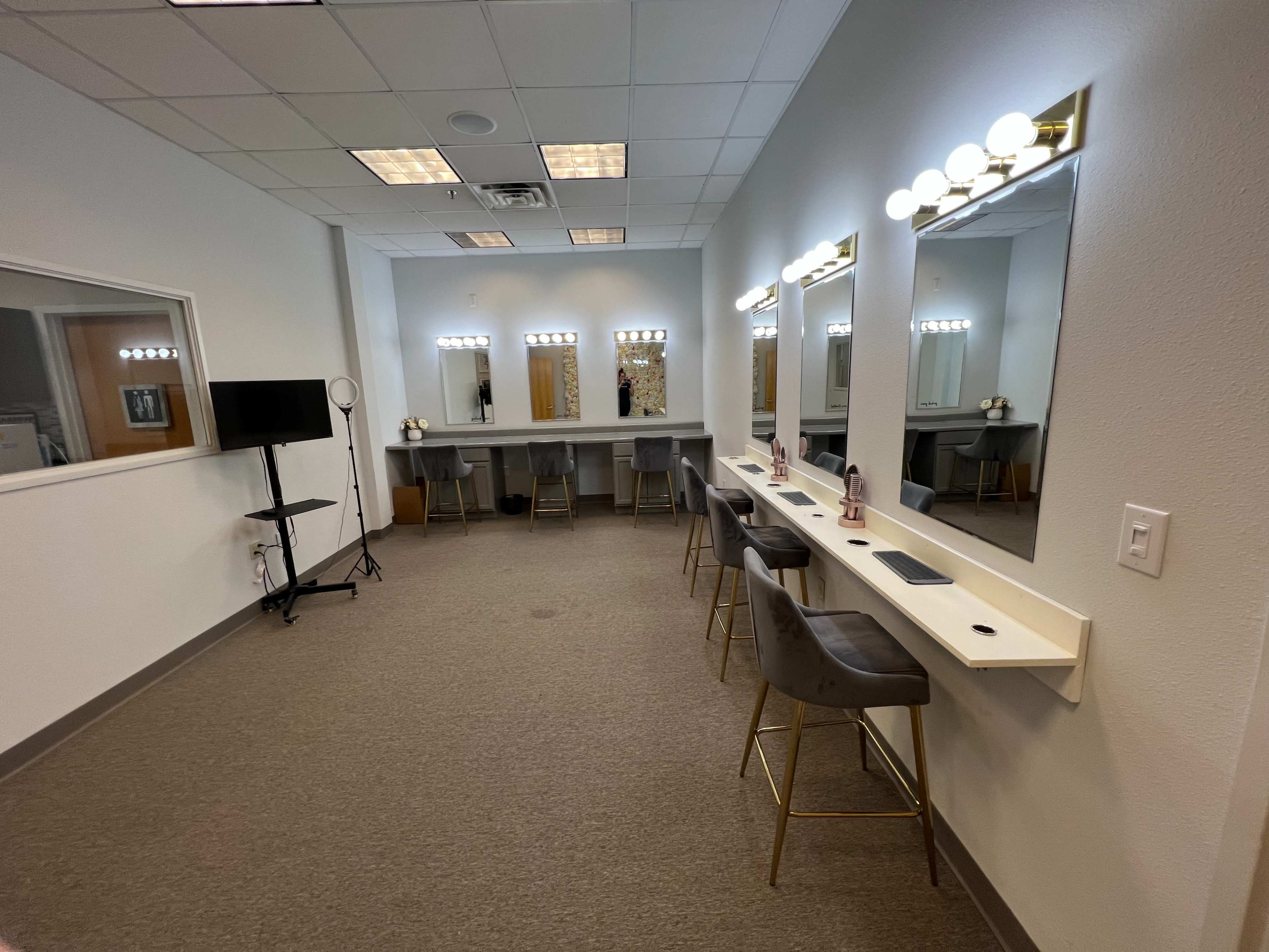 A well-lit room featuring three mirrors with lights, a long countertop with four stools, and a stand for a microphone or light on the side.