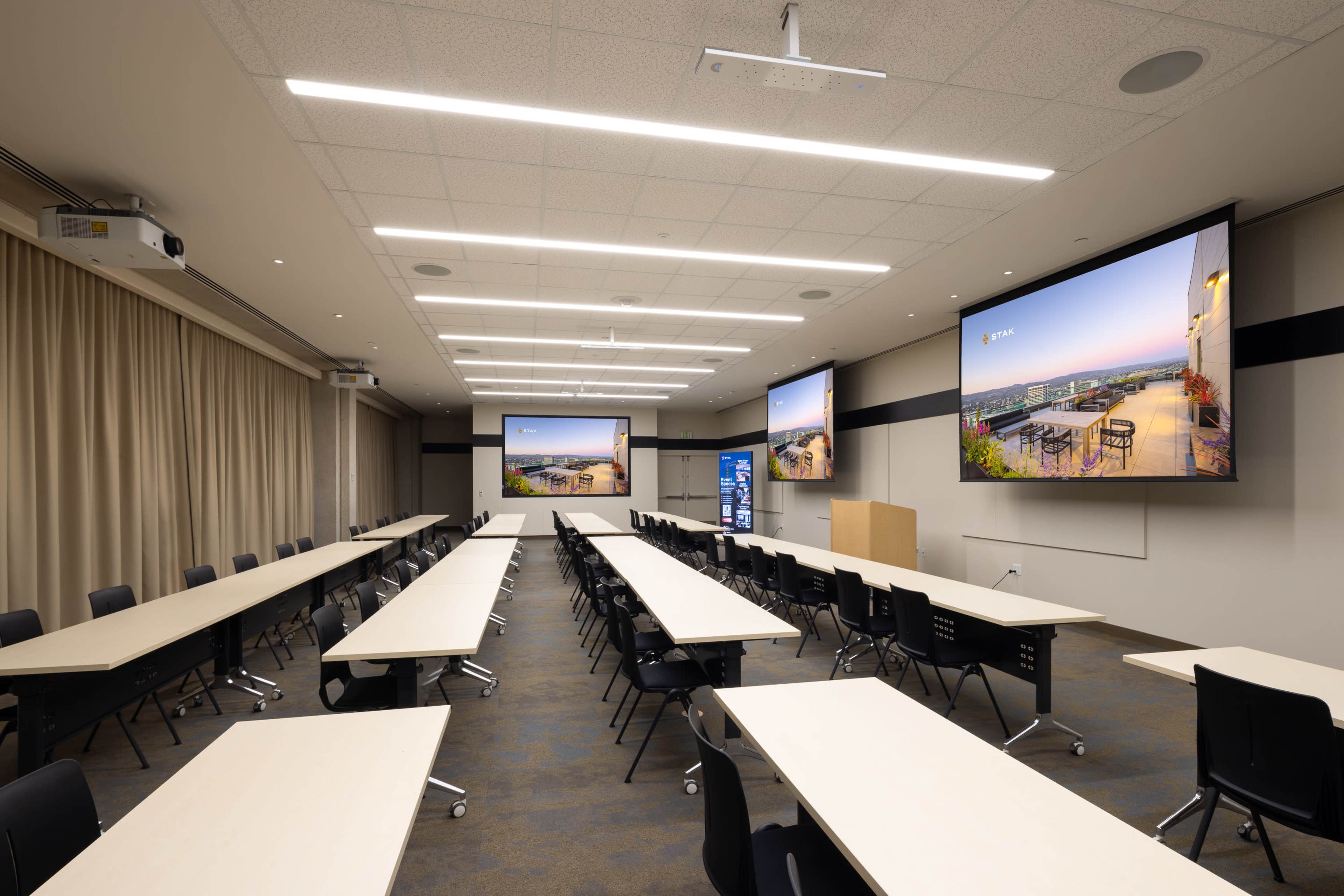 A modern conference room features two large screens at the front and rows of tables and chairs arranged in a flexible layout.