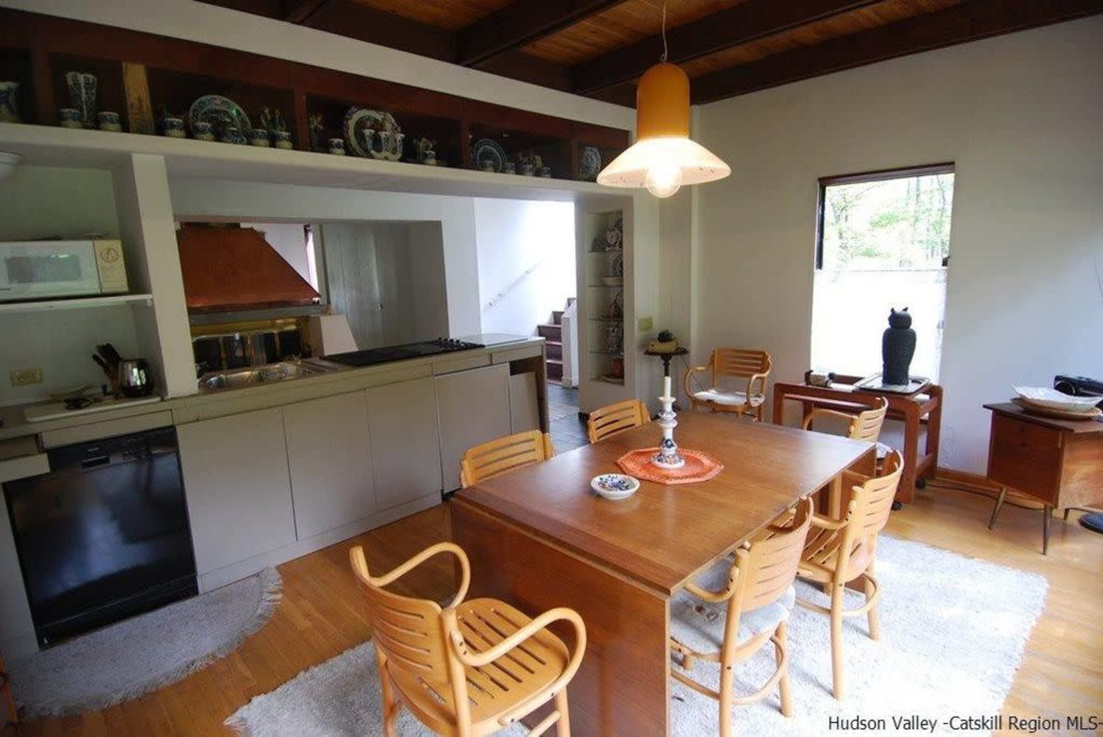 The image shows a kitchen and dining area featuring a wooden table surrounded by chairs, with a small kitchenette in the background.