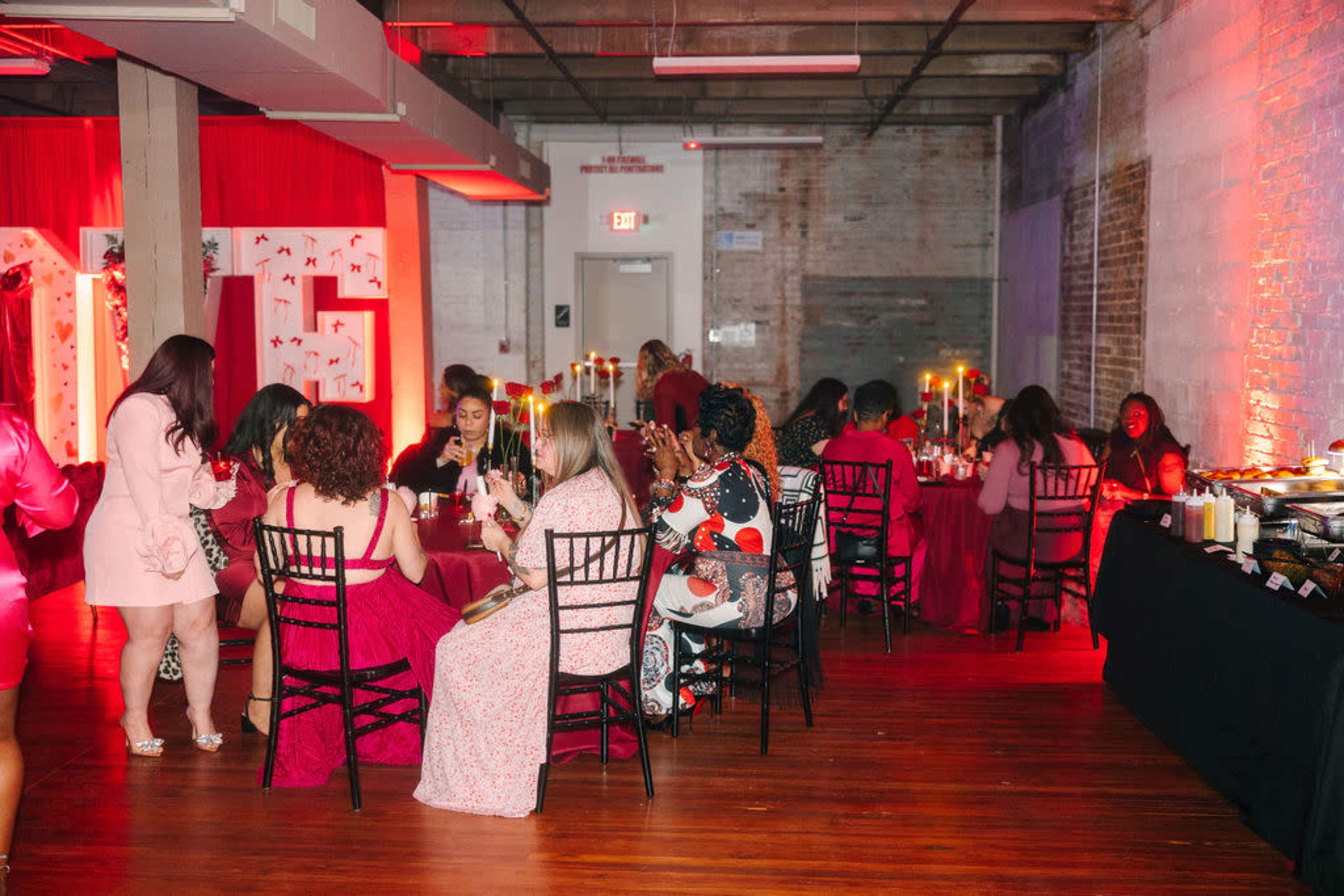 The image shows a gathering with guests seated at tables, decorated in red and pink tones, enjoying food and drinks in an ornate room with dim lighting.