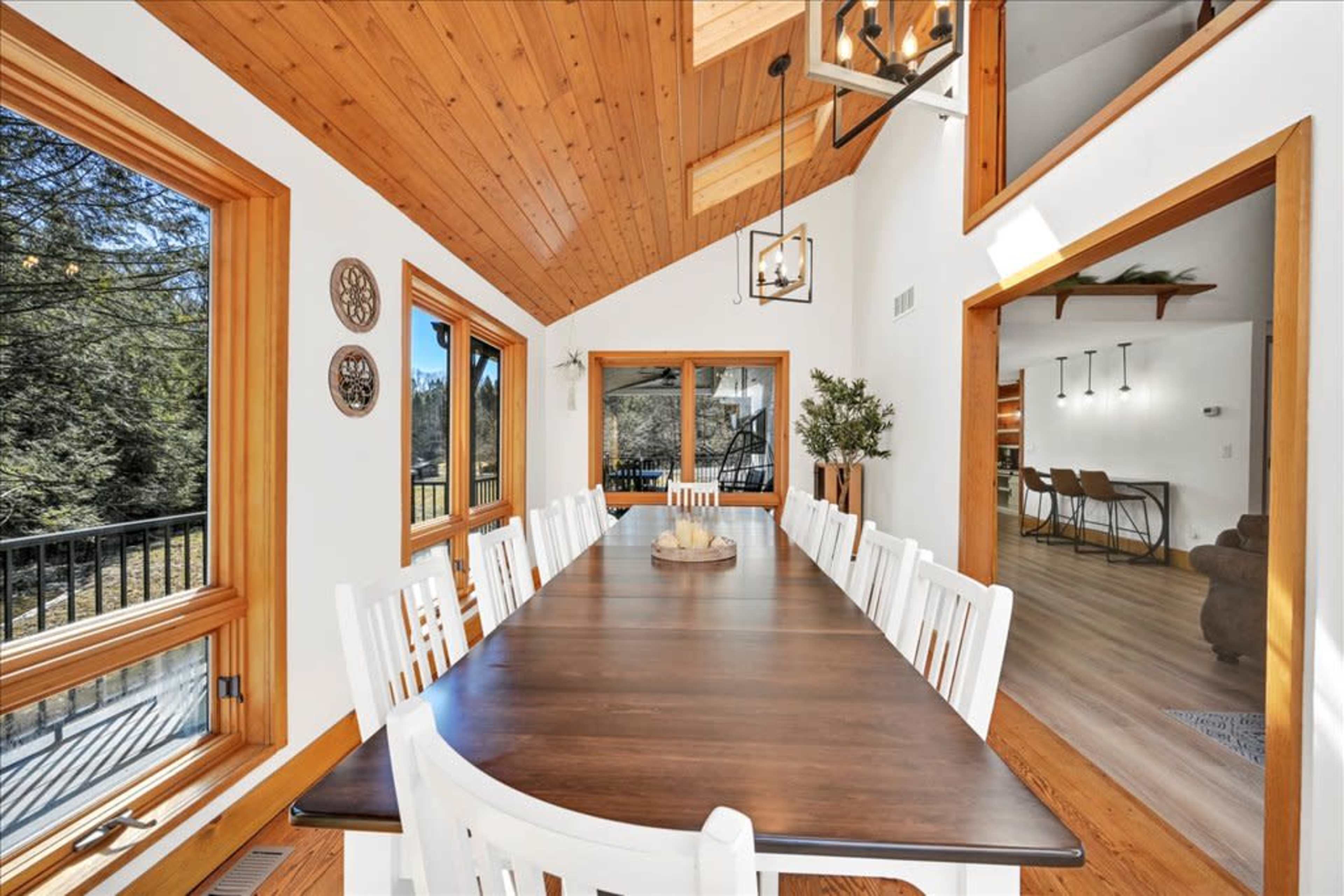A spacious dining area features a long wooden table surrounded by white chairs, with large windows allowing natural light to illuminate the room.