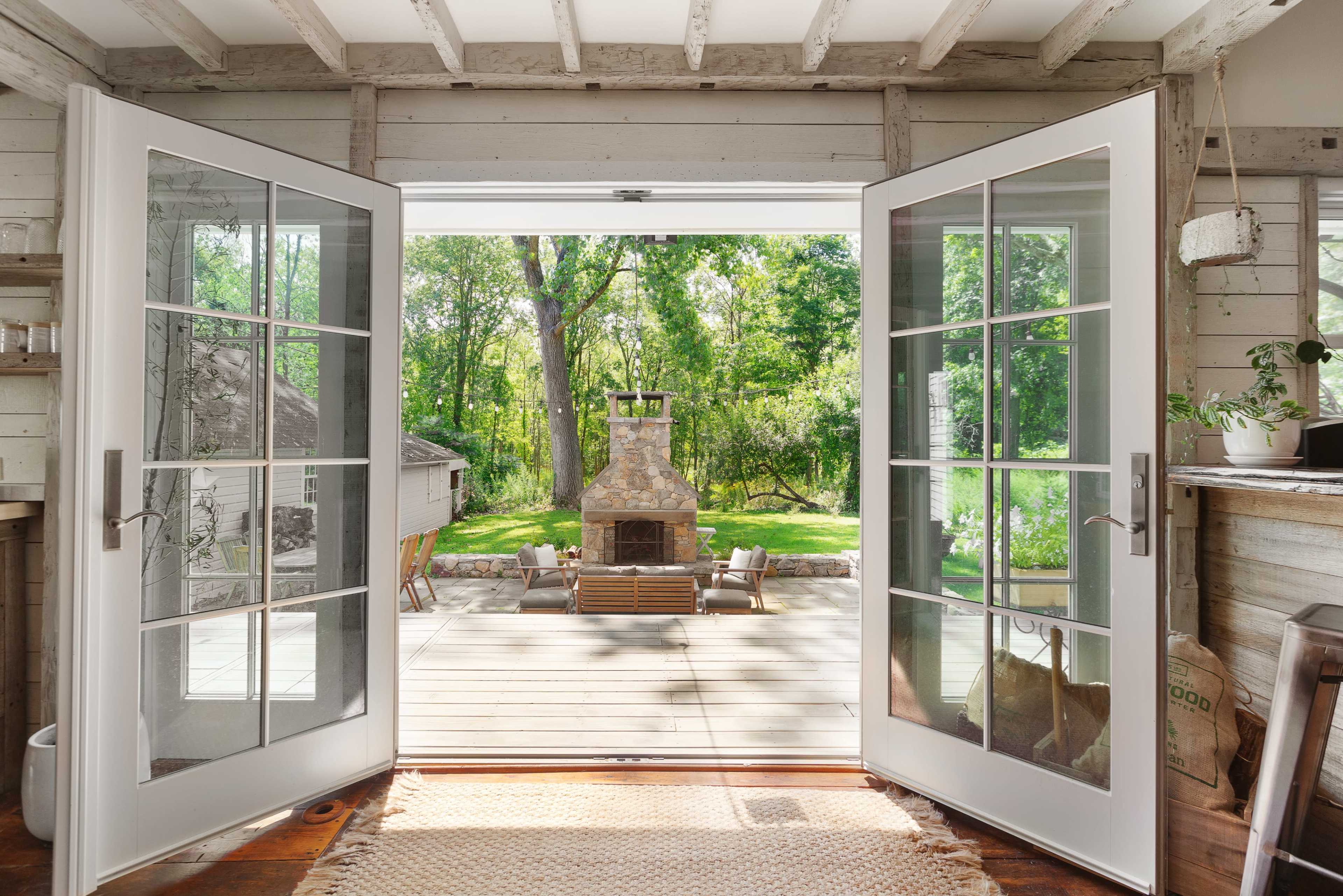 A pair of open French doors leads to a deck with a stone fireplace and chairs set against a backdrop of greenery.