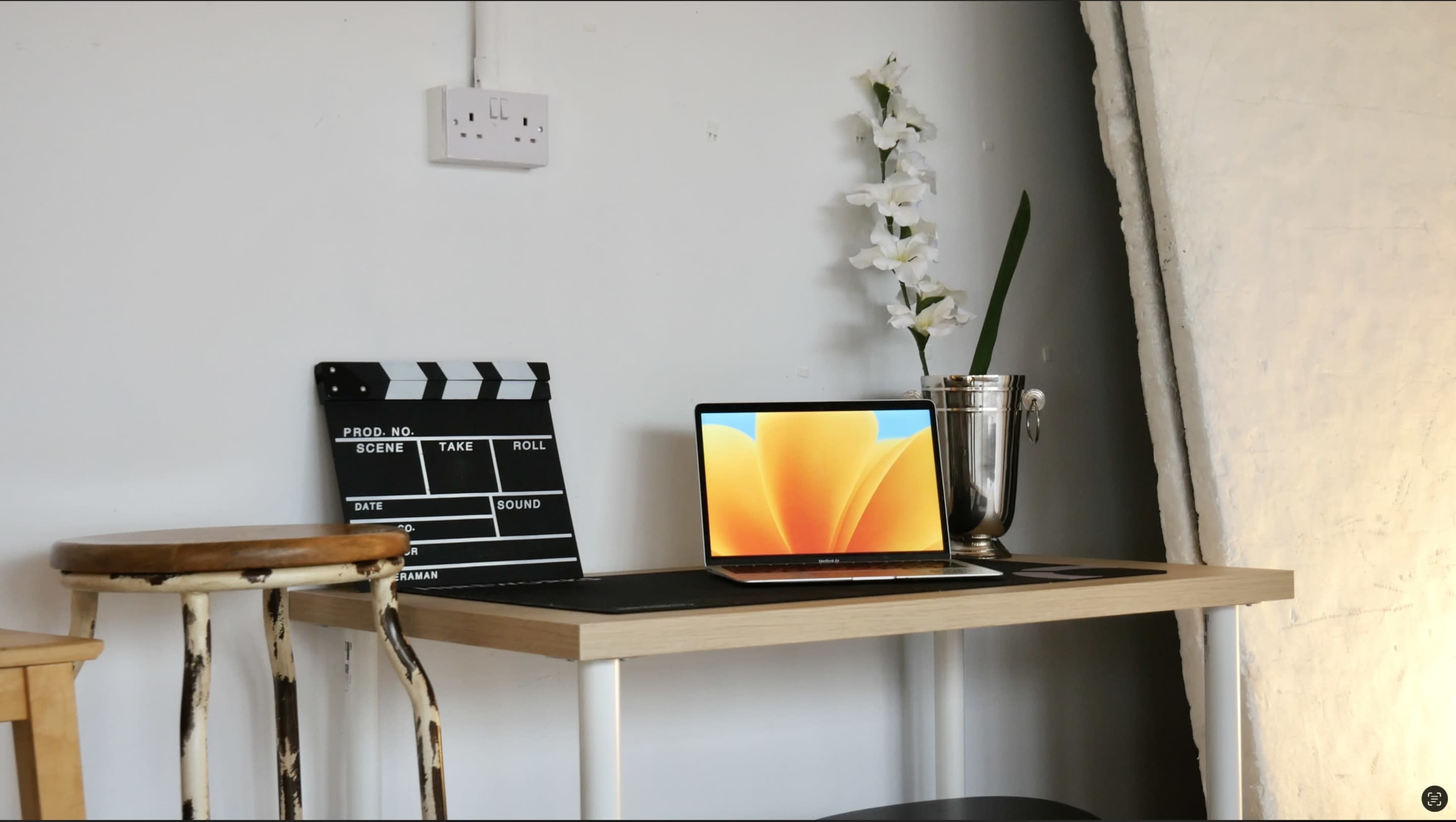 A minimalist desk features a laptop, a movie clapboard, a silver vase with flowers, and a bar stool against a simple wall backdrop.