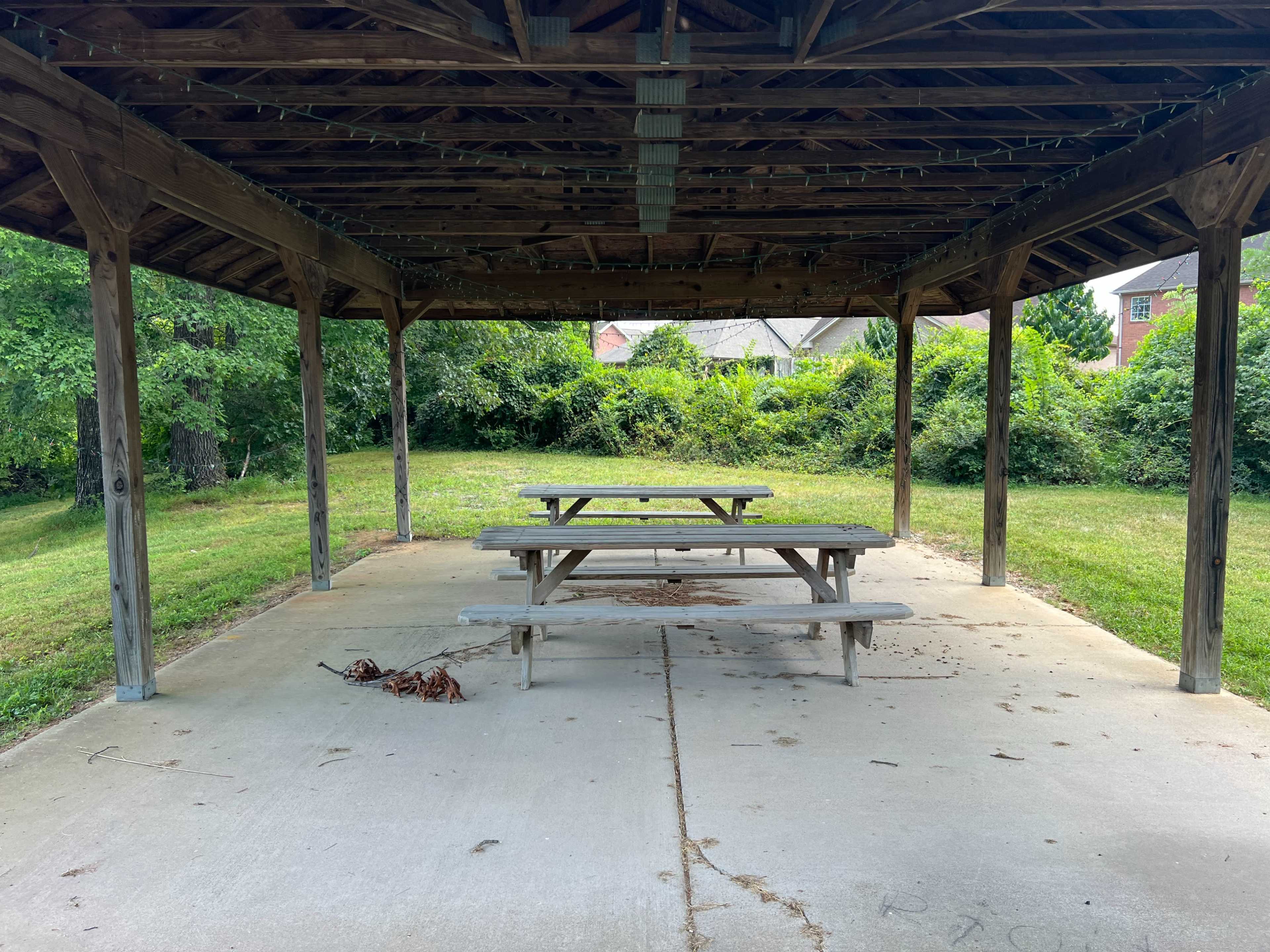 The image shows a grassy area with a wooden pavilion featuring several picnic tables beneath its roof.