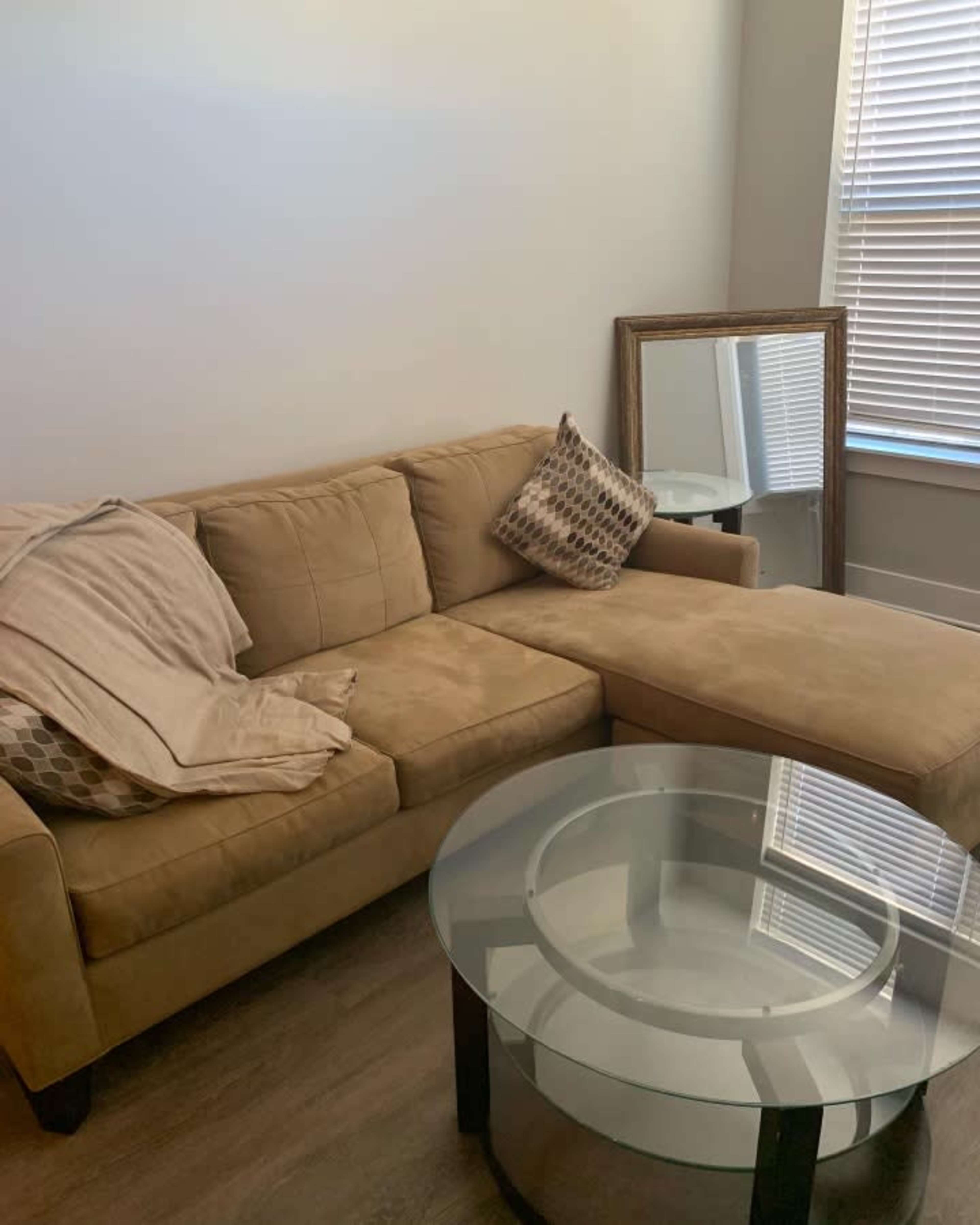 A beige sectional sofa with a decorative pillow sits beside a round glass coffee table and a mirror reflecting natural light from a nearby window.