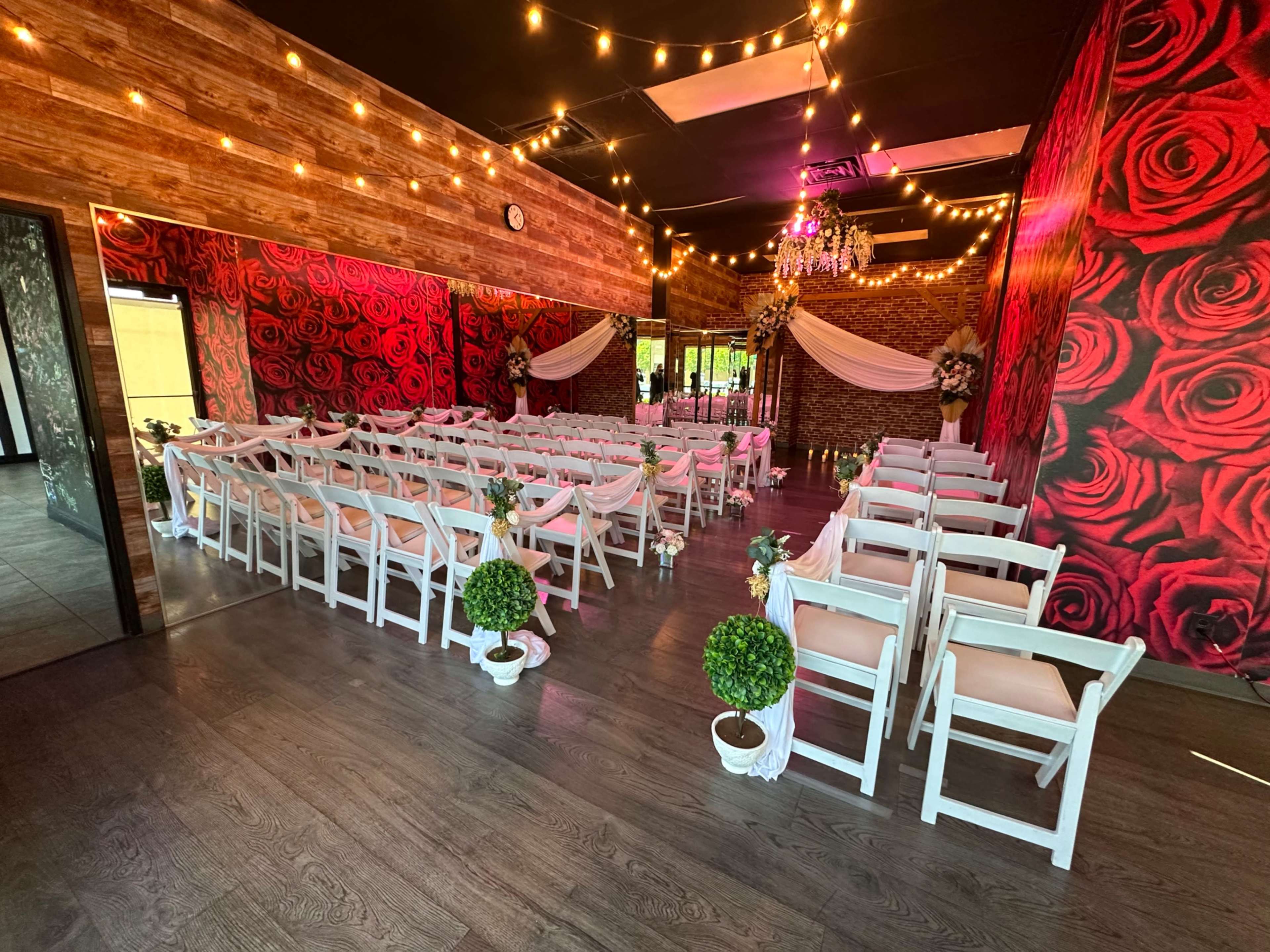 A wedding venue with rows of white chairs arranged in front of a floral backdrop adorned with roses and decorative lighting.