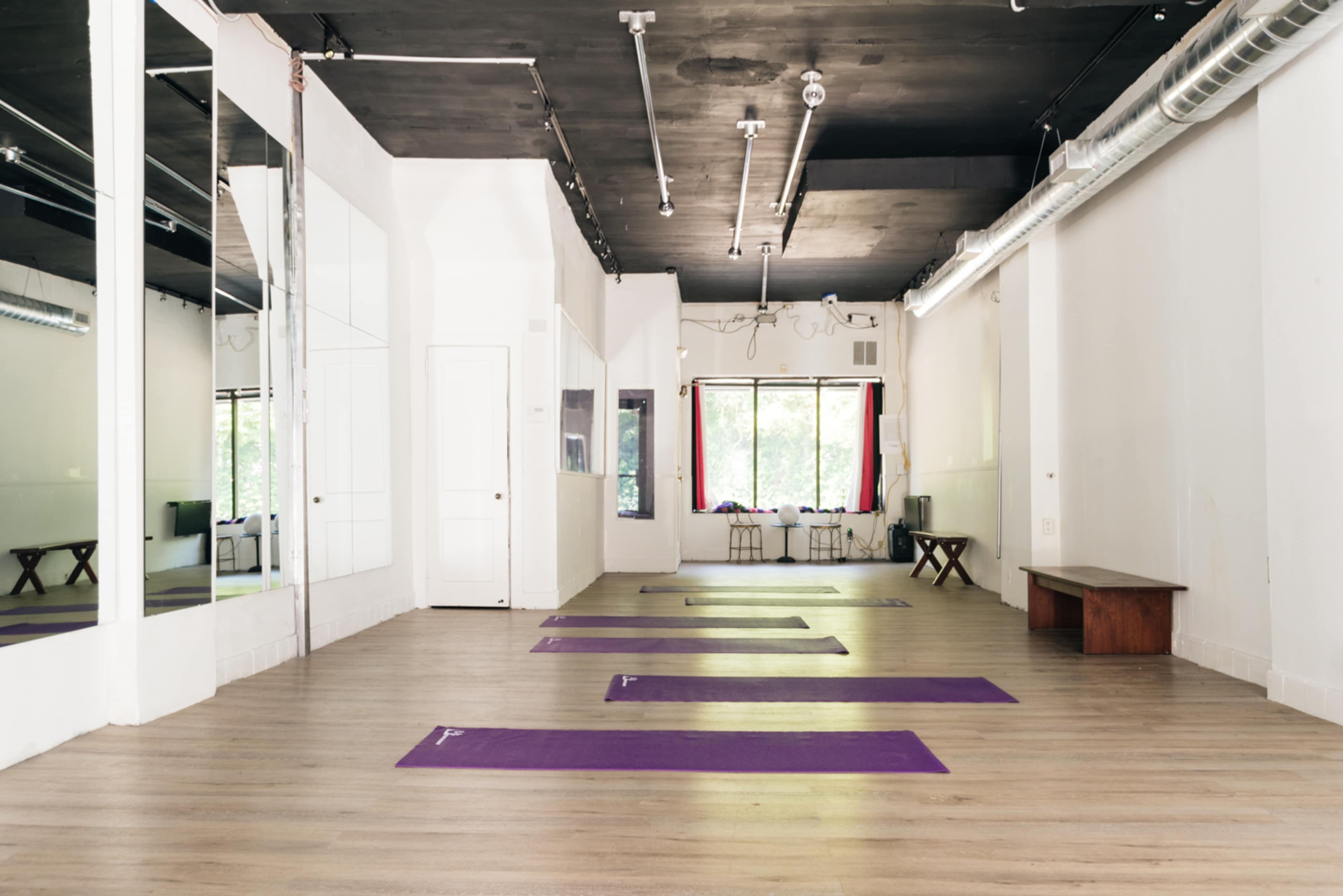 A spacious yoga studio features several purple mats arranged on a light wooden floor, with large windows providing natural light.