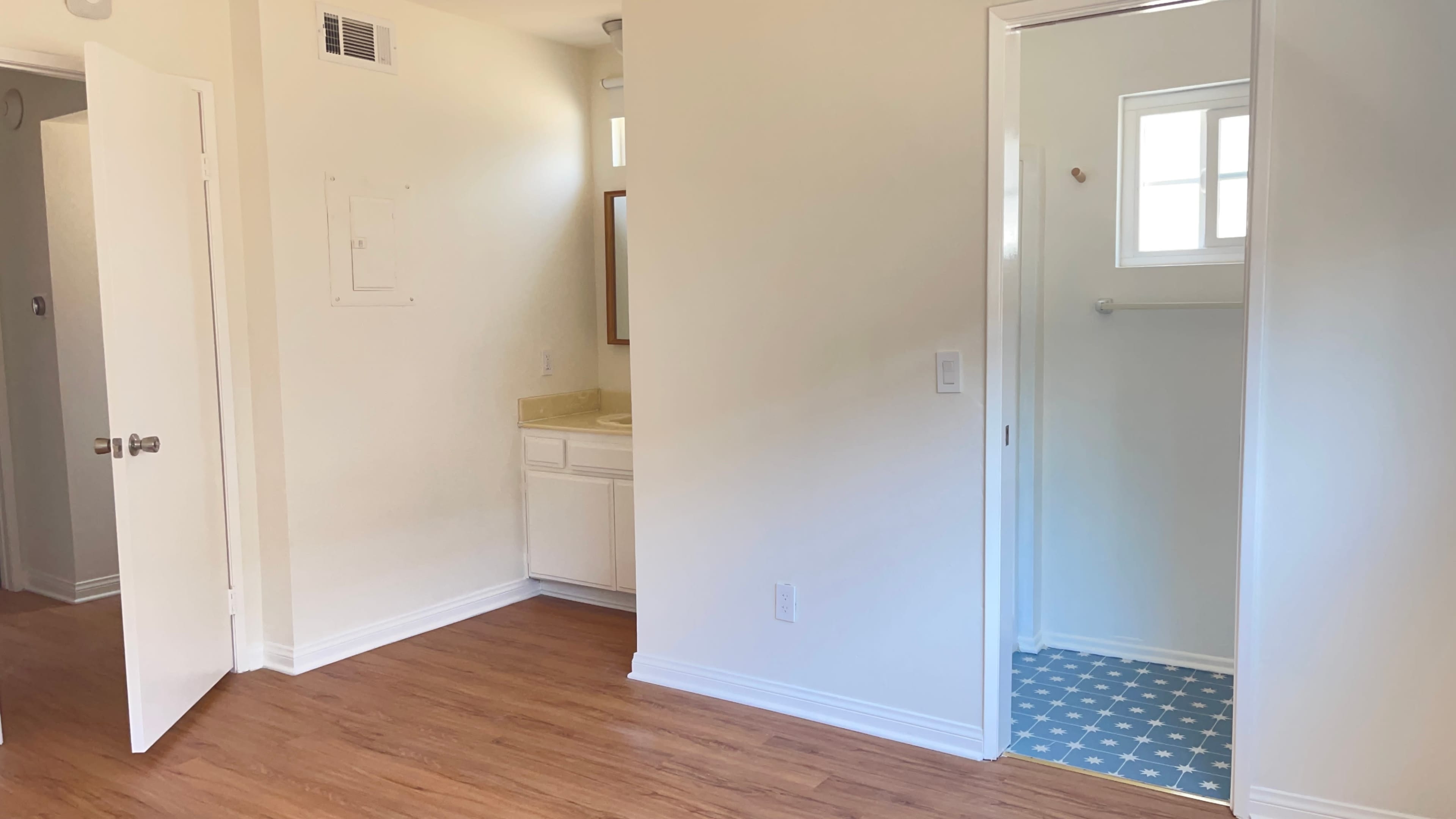 A room with wooden flooring, a doorway leading to a bathroom with a tiled floor, and a space for a vanity with a mirror on the wall.