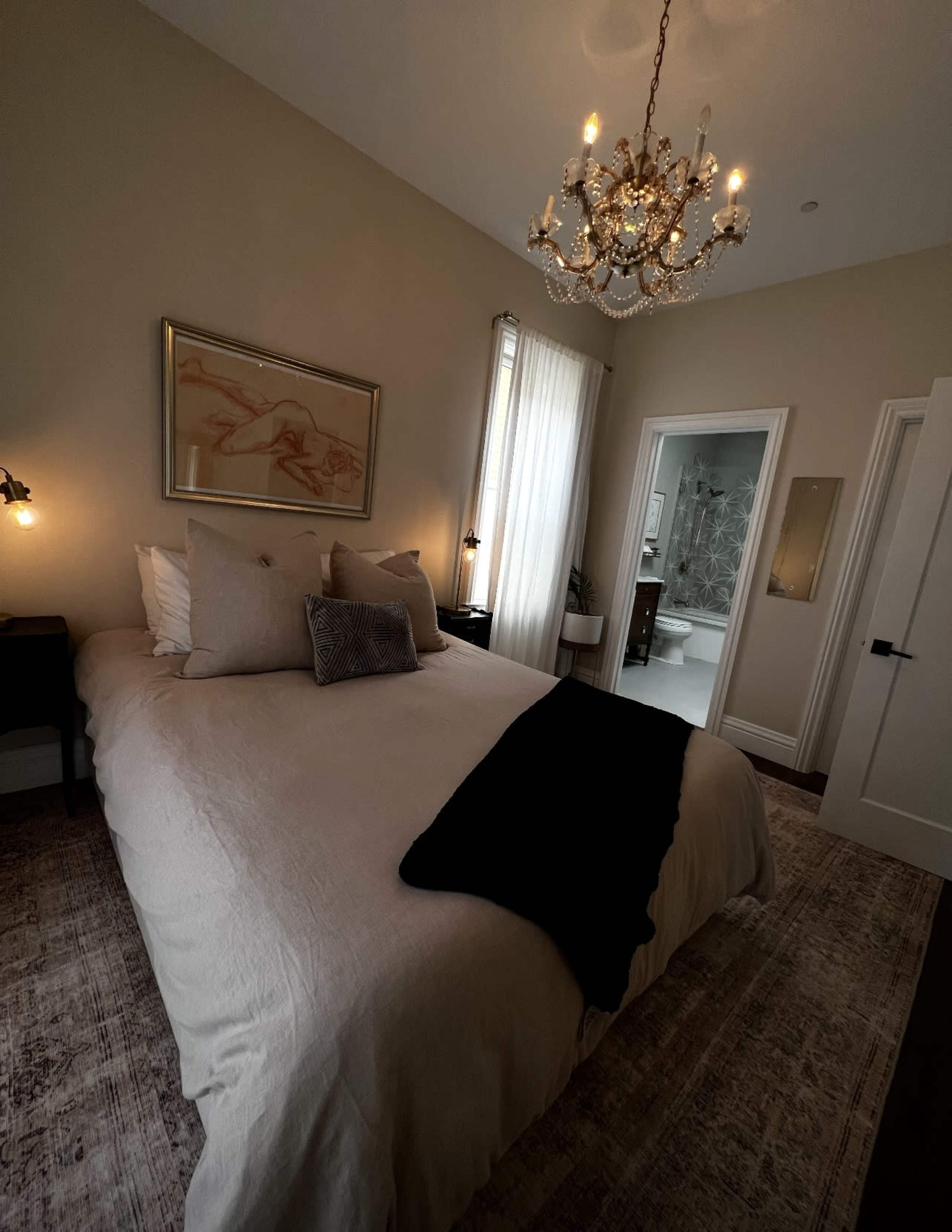 A softly lit bedroom features a large bed with neutral bedding, a chandelier overhead, and an adjoining bathroom visible through an open doorway.