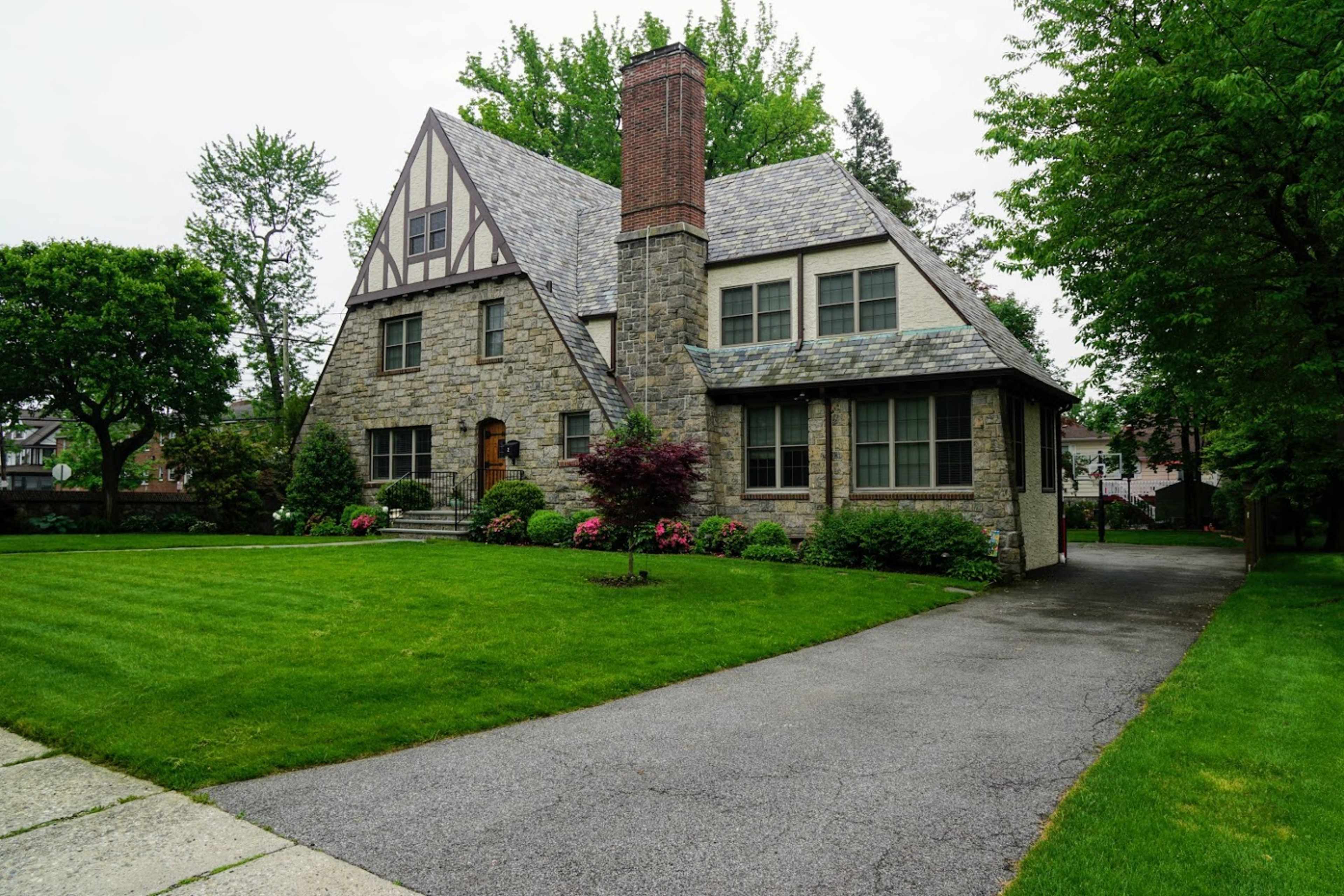 The image shows a large, stone Tudor-style house with a slate roof, situated on a manicured lawn alongside a paved driveway.