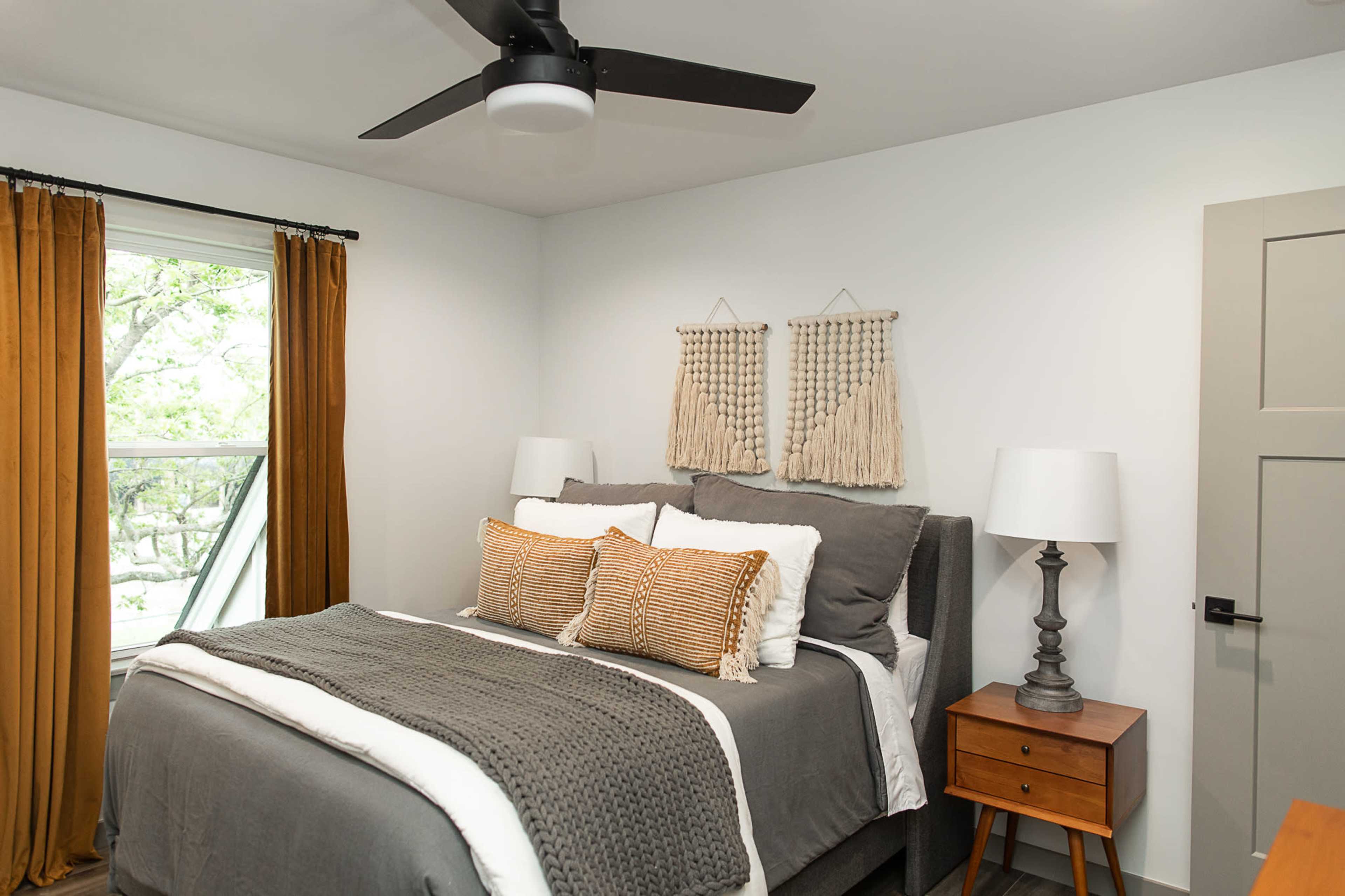 A modern bedroom featuring a gray bed with decorative pillows, a bedside table with a lamp, and large windows with yellow curtains.
