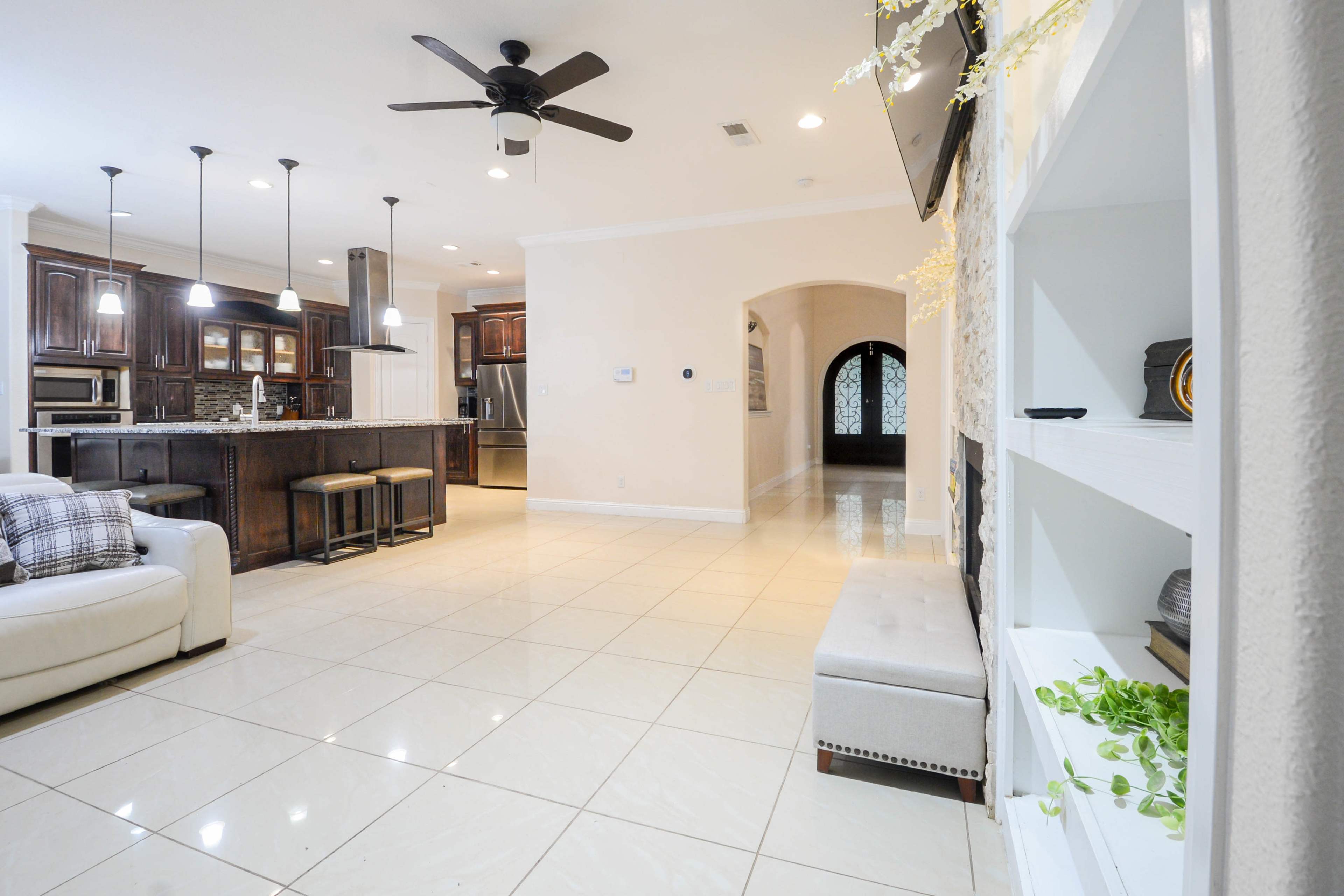 The image shows a spacious living area with modern furniture, a large kitchen in the background, and an archway leading to another room.