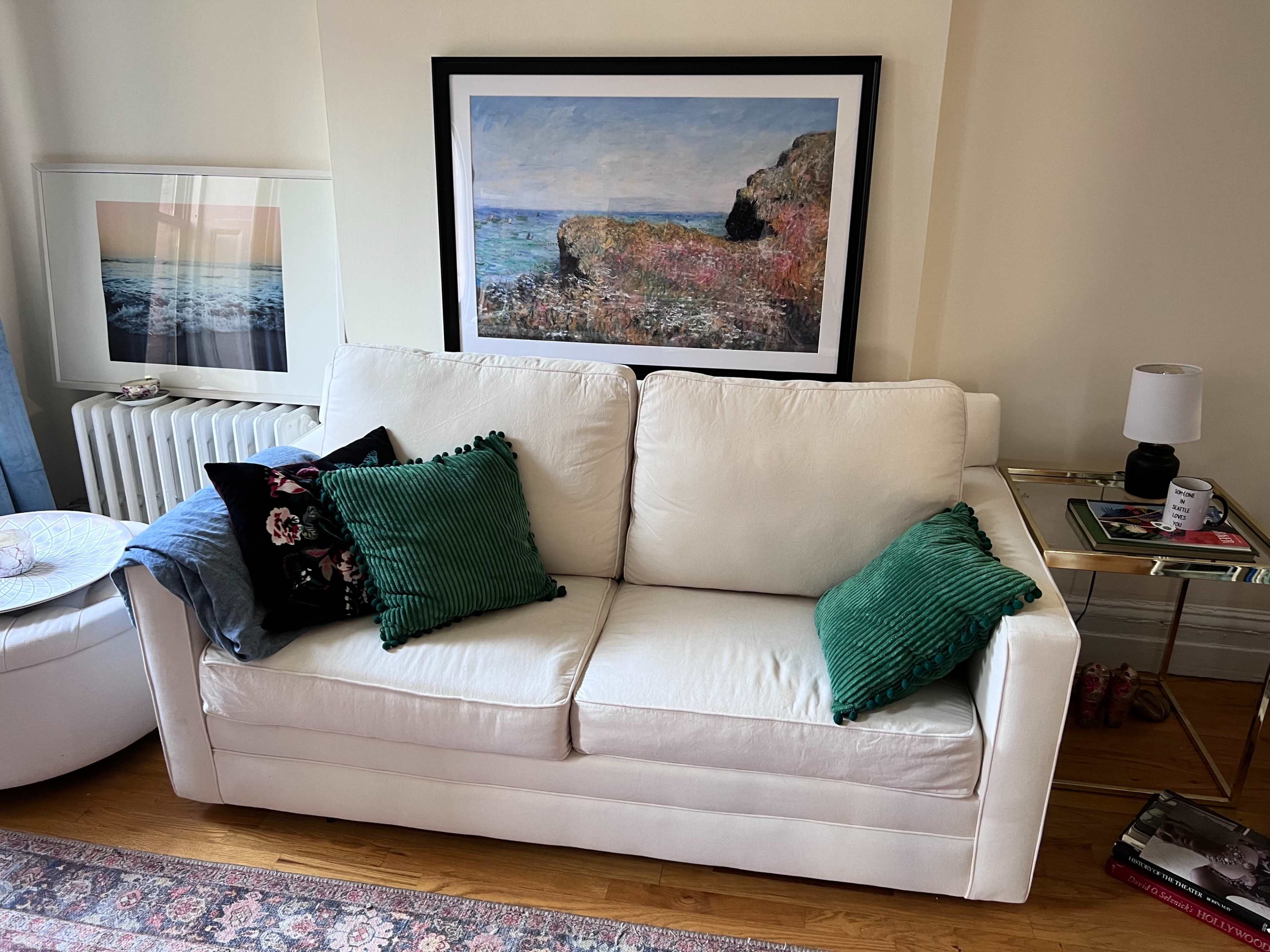 A white sofa with green and floral-patterned pillows sits in a room adorned with art and a small side table.
