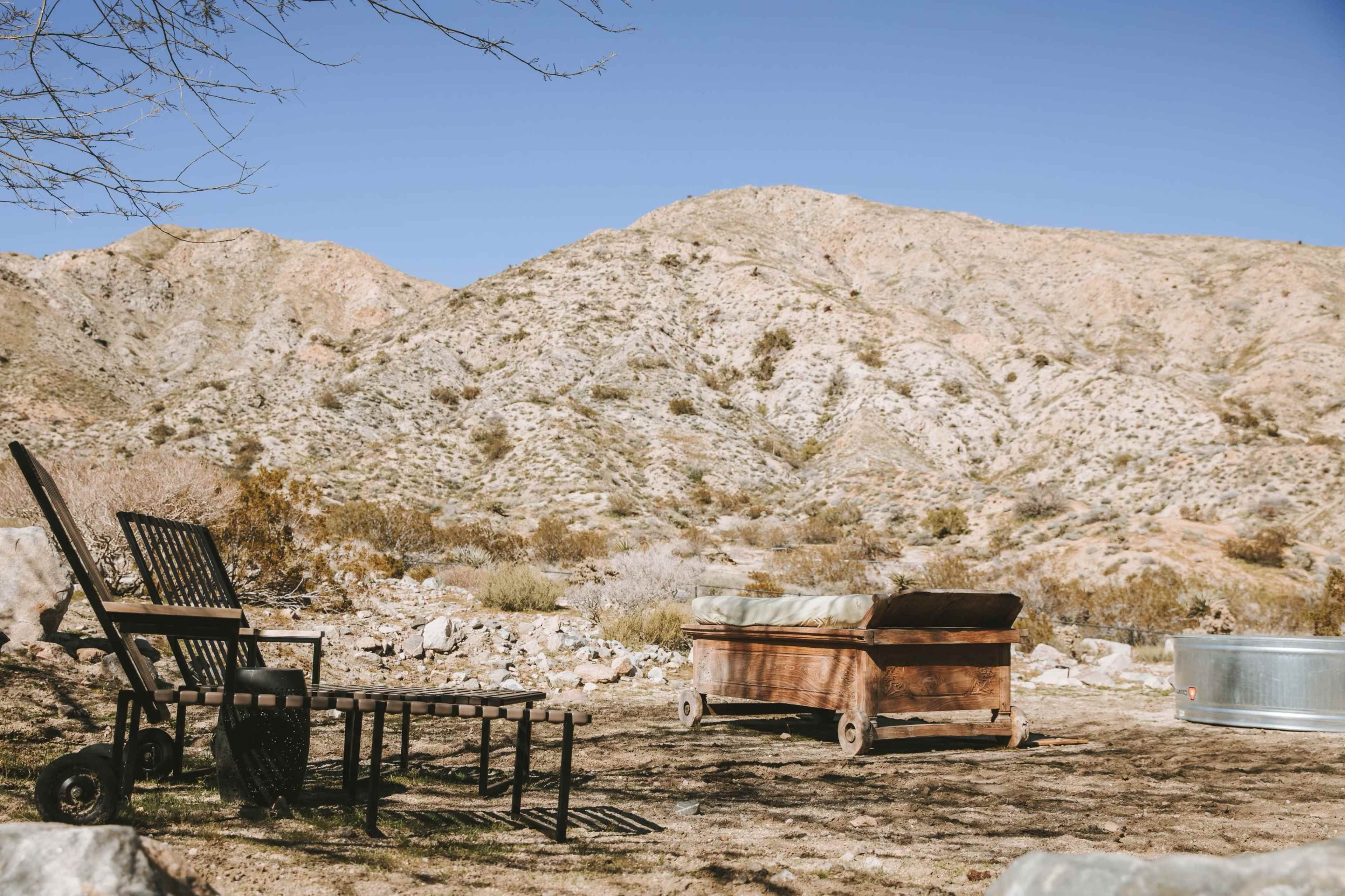 The scene shows outdoor furniture, including a chair and a wooden couch, set against a backdrop of rocky, desert hills under a clear blue sky.