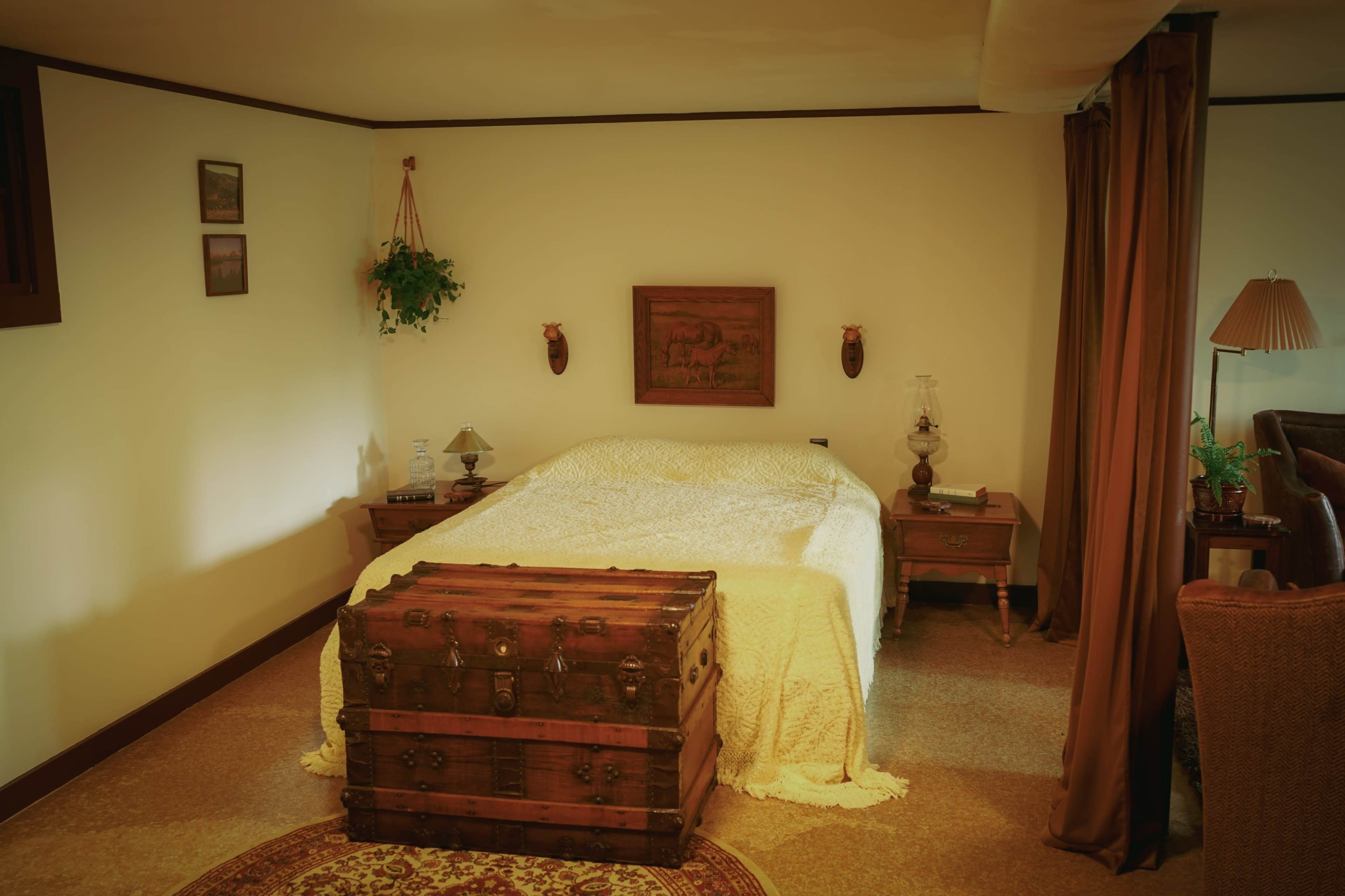 The image shows a cozy bedroom featuring a bed with a white coverlet, flanked by two bedside tables, and a decorative trunk at the foot of the bed.