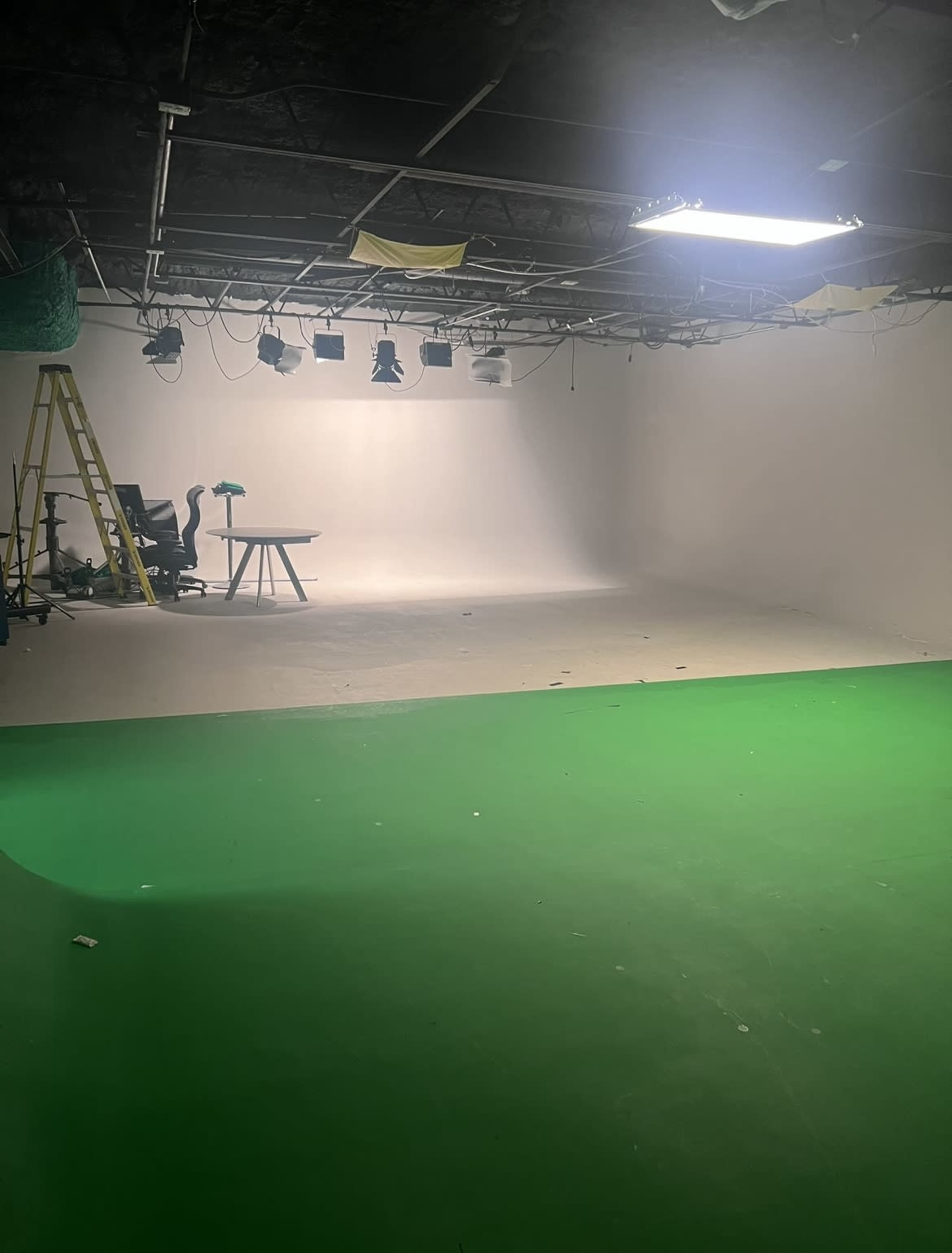 A partially equipped studio space with a green floor, lighting fixtures hanging from the ceiling, and a ladder leaning against the wall.