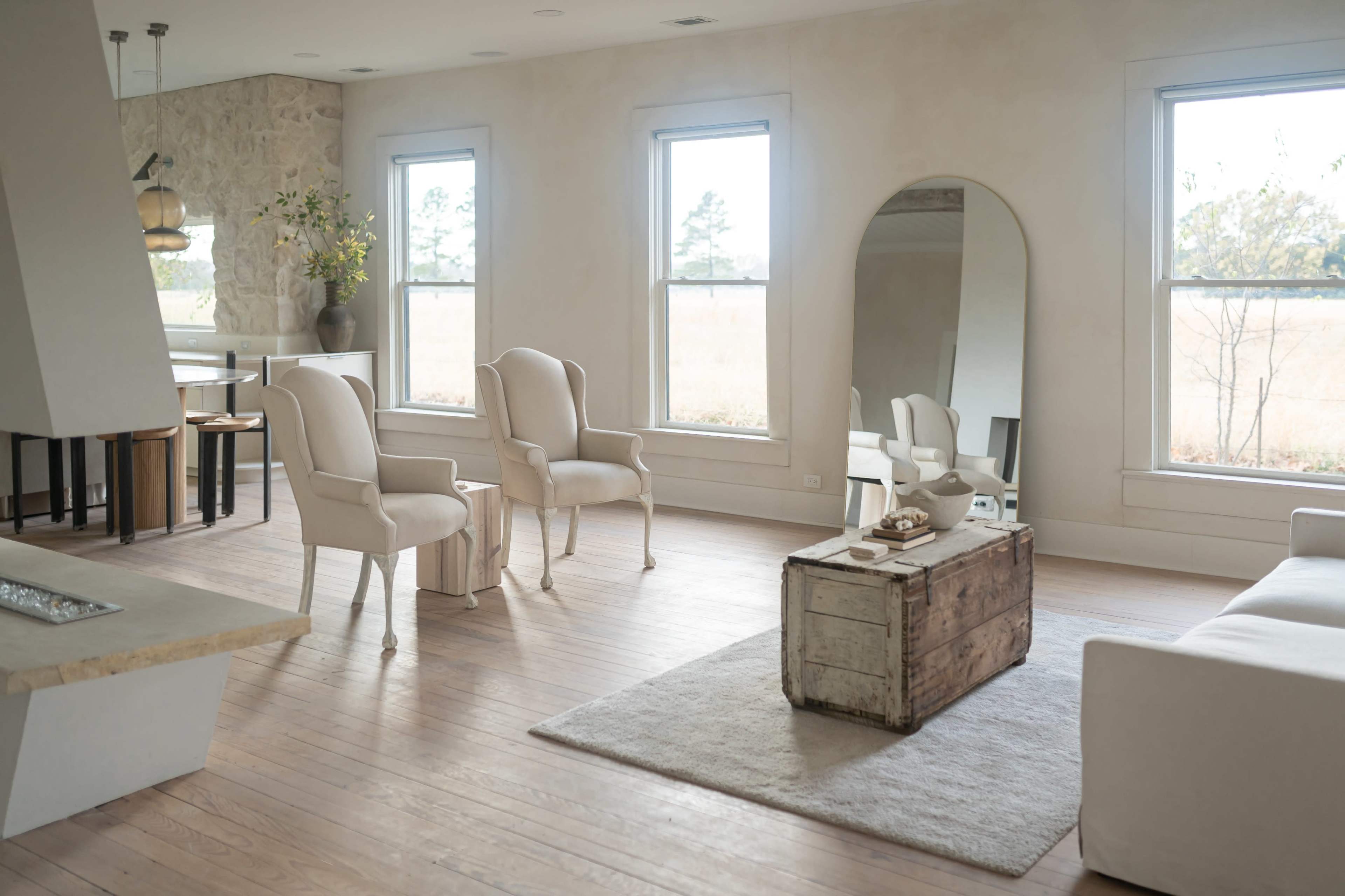 The image depicts a spacious, light-filled interior featuring two upholstered chairs, a rustic wooden coffee table, and large windows that overlook an outdoor view.