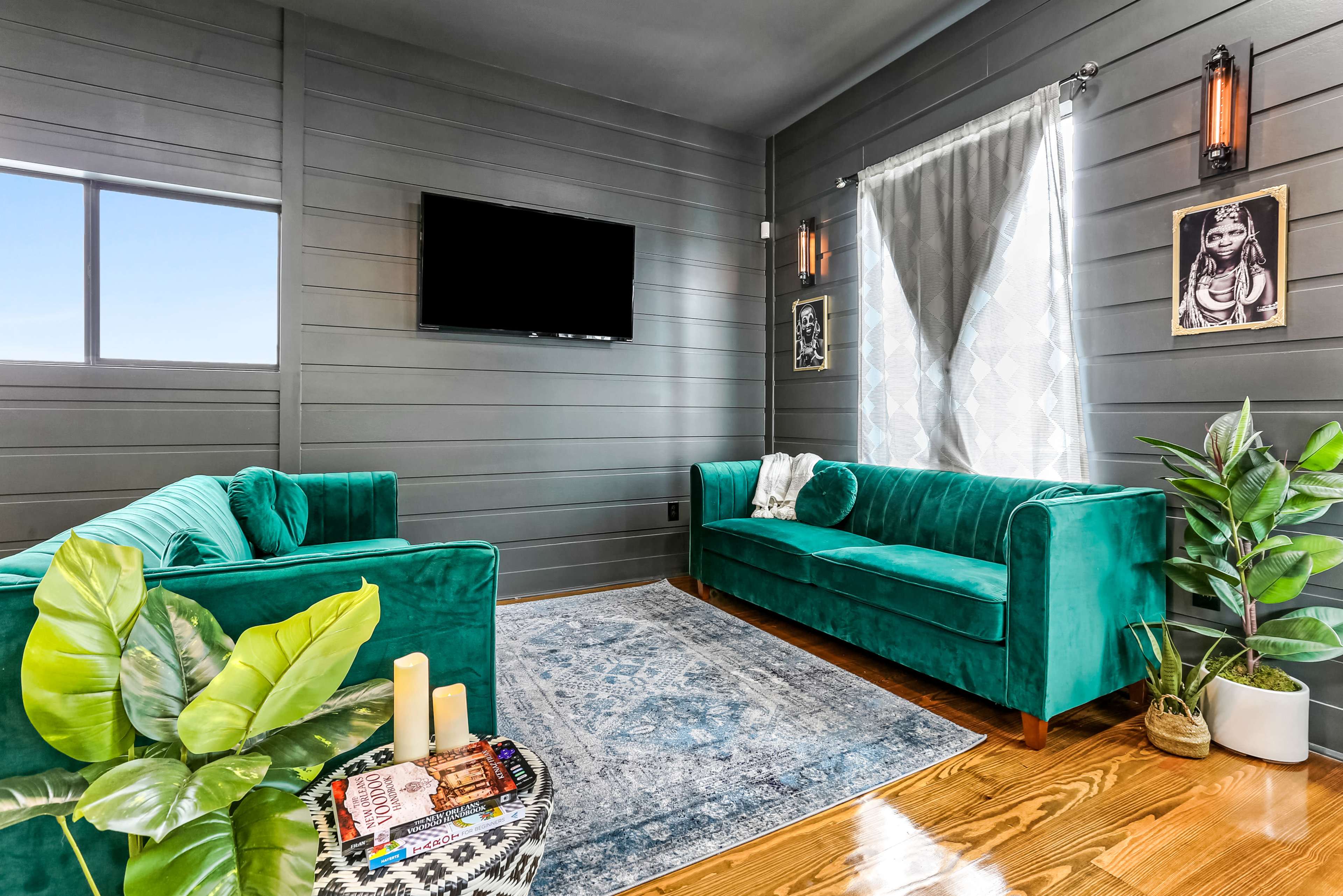 The room features two teal sofas, a patterned rug, a television mounted on the wall, and a large window with sheer curtains.