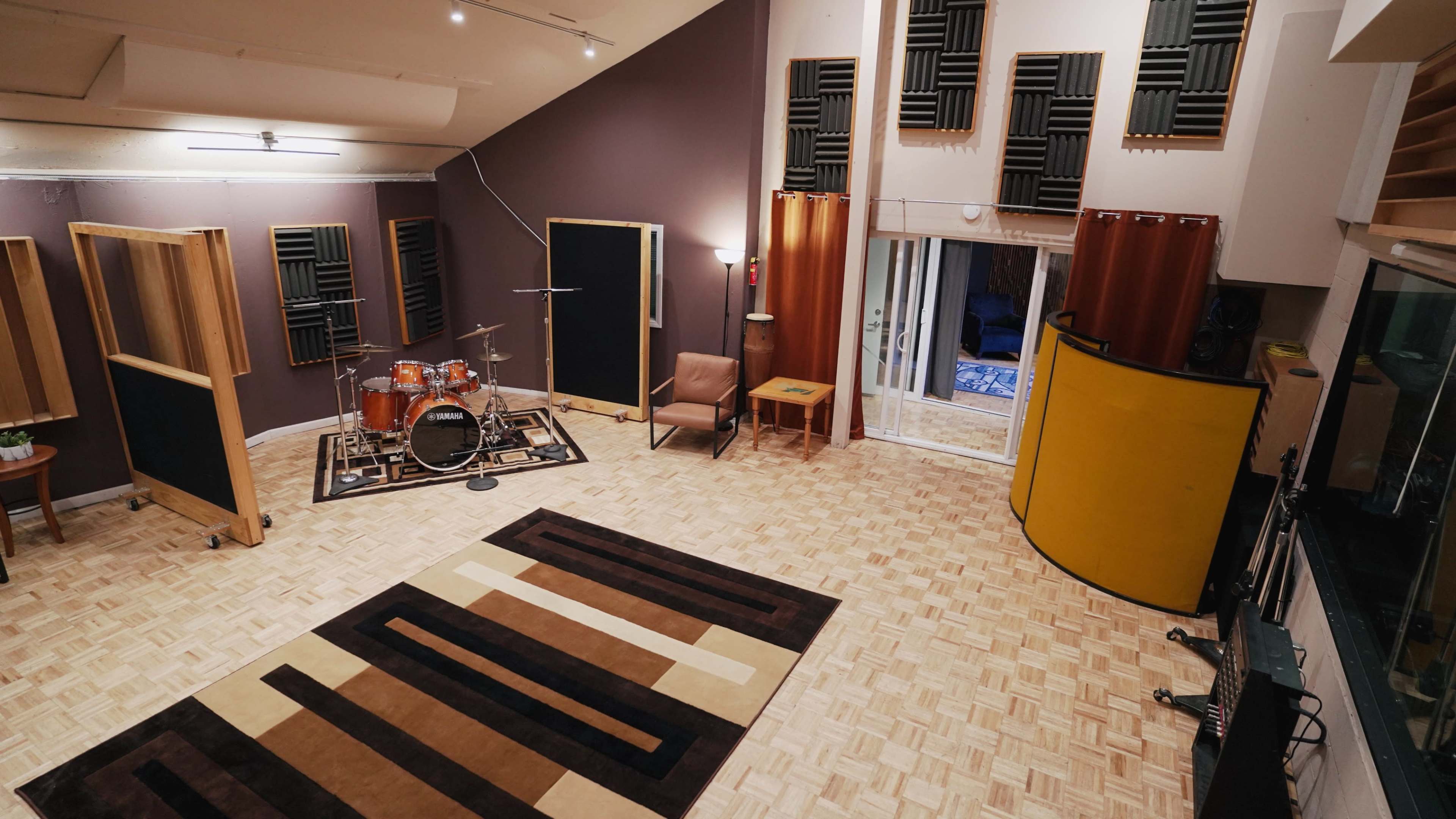 The image shows a music recording studio featuring a drum set, acoustic panels on the walls, and a patterned rug on the wooden floor.