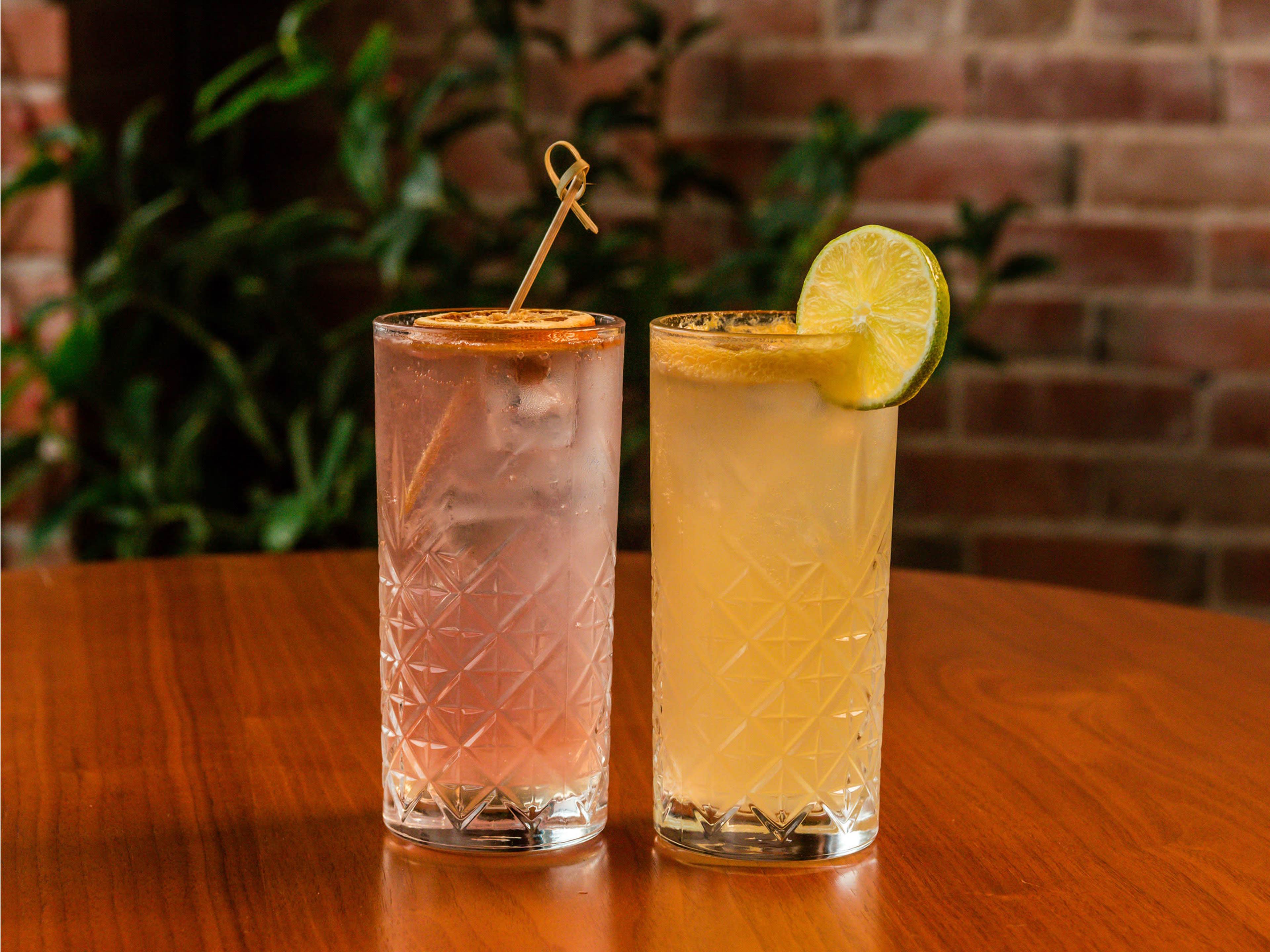 Two tall, clear glasses filled with colorful beverages are positioned on a wooden table, with one drink garnished with a lime slice and the other with a straw.