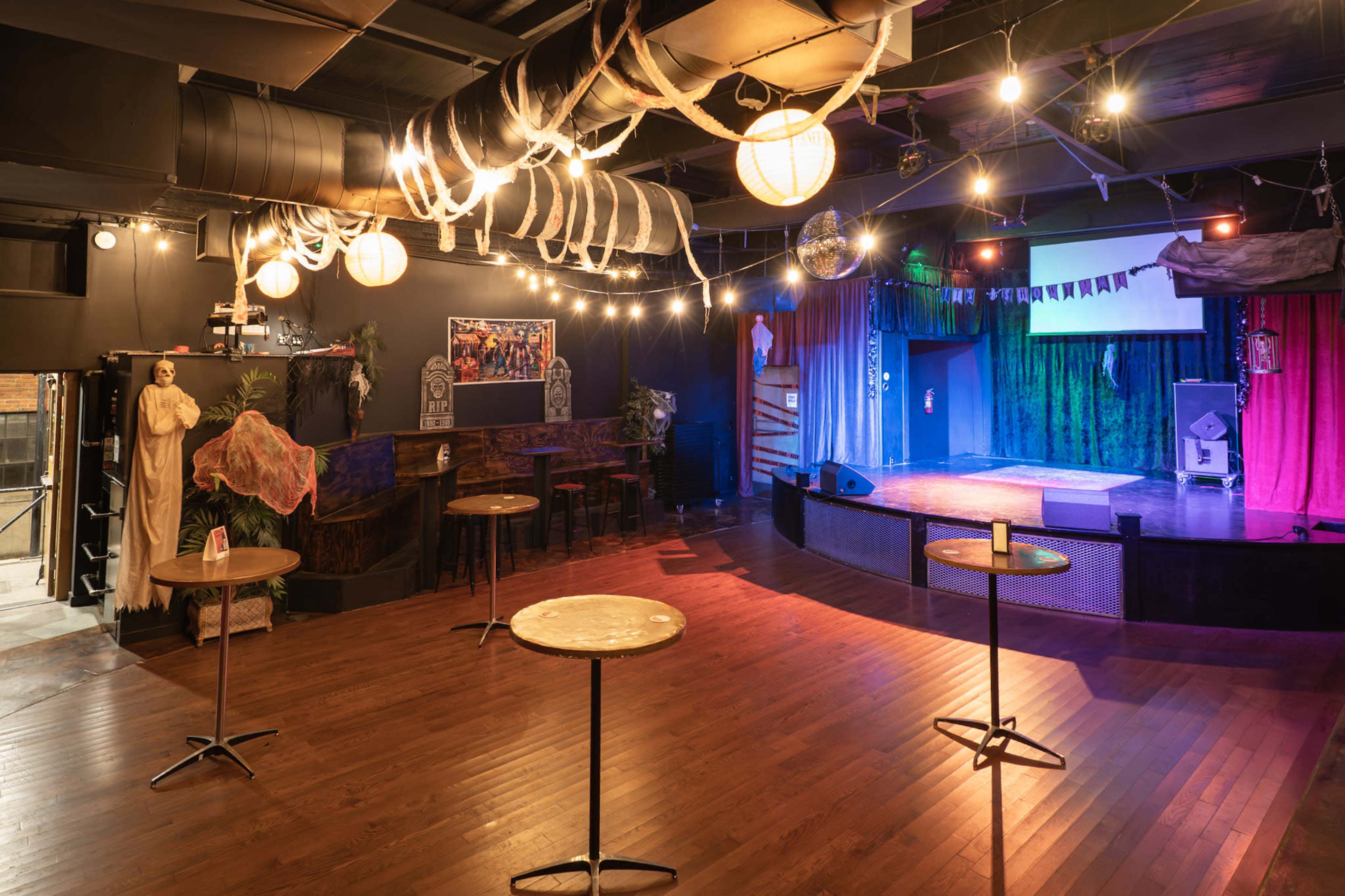 The image shows an empty performance venue with a stage at the front and several small tables scattered around, illuminated by hanging lights.