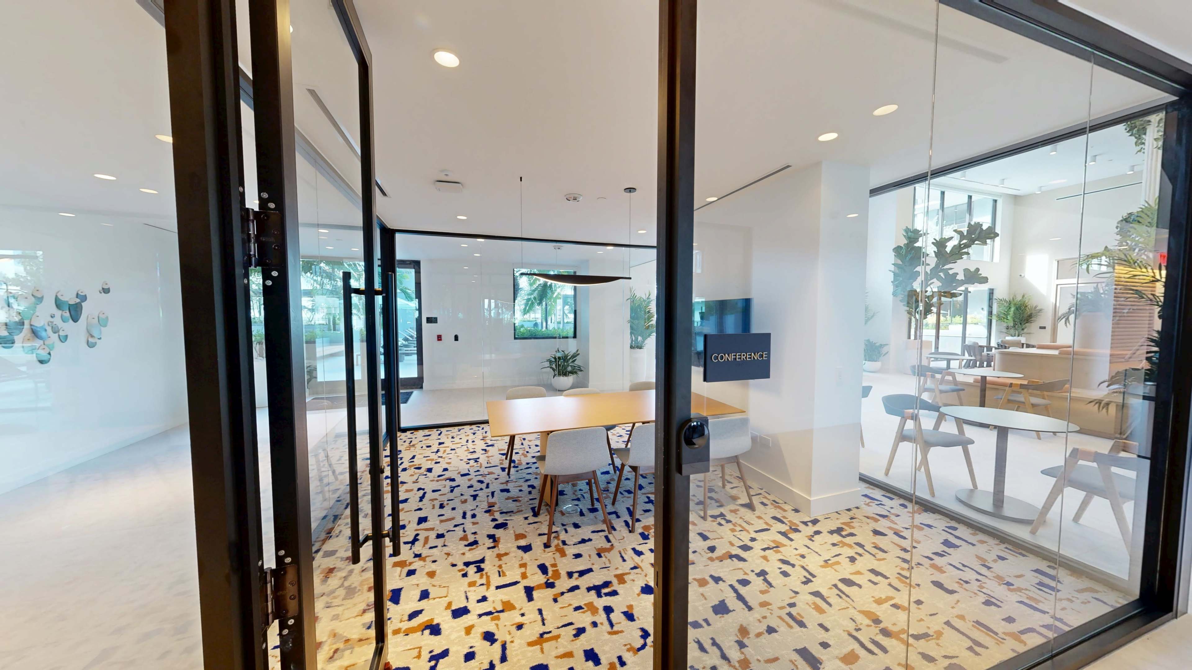The image shows a modern conference room with a large table, chairs, and a wall-mounted screen, surrounded by glass doors and decorated with plants.