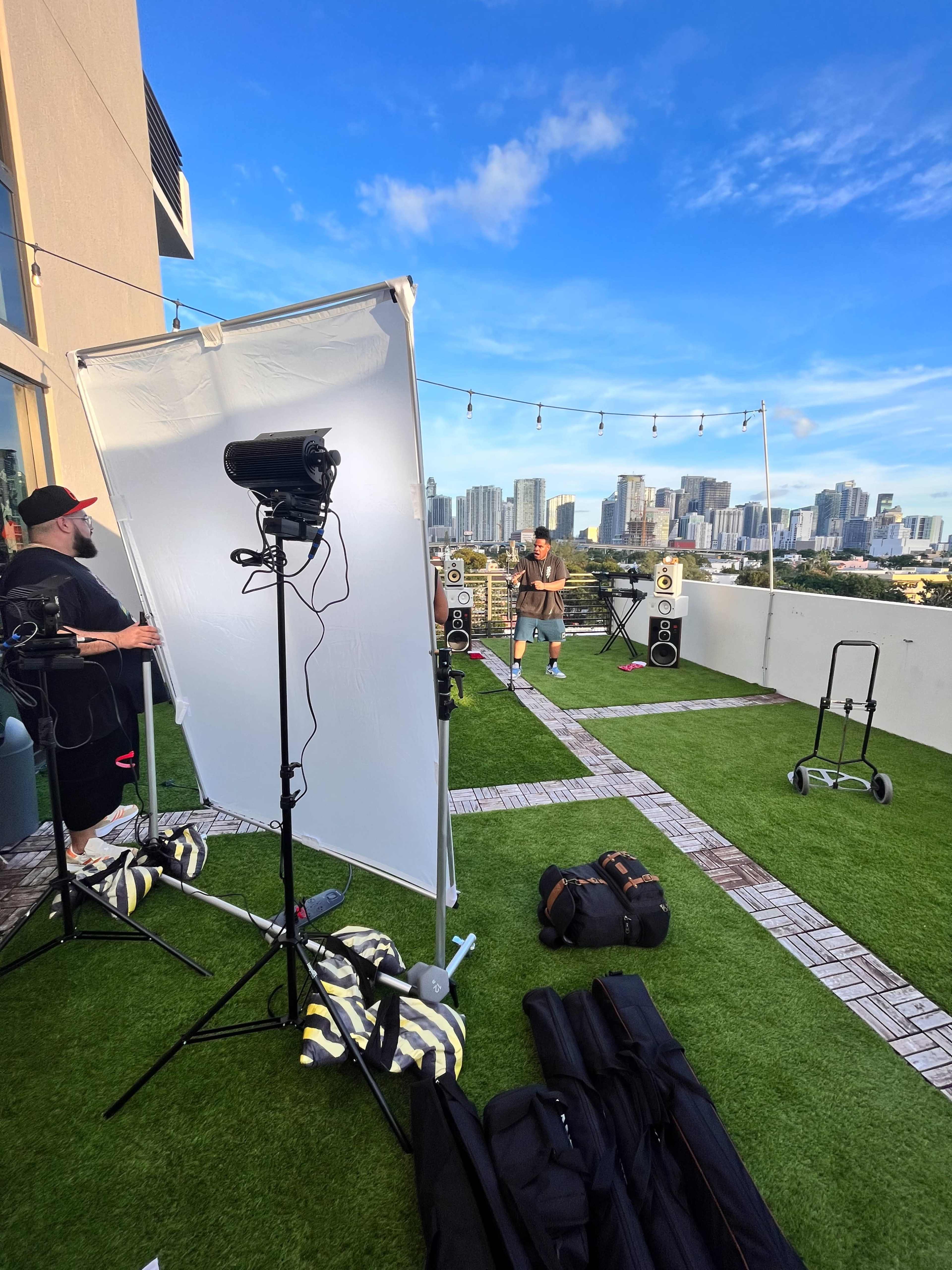 A man stands on a rooftop patio setting with city skyscrapers in the background, while two crew members adjust lighting equipment and speakers.
