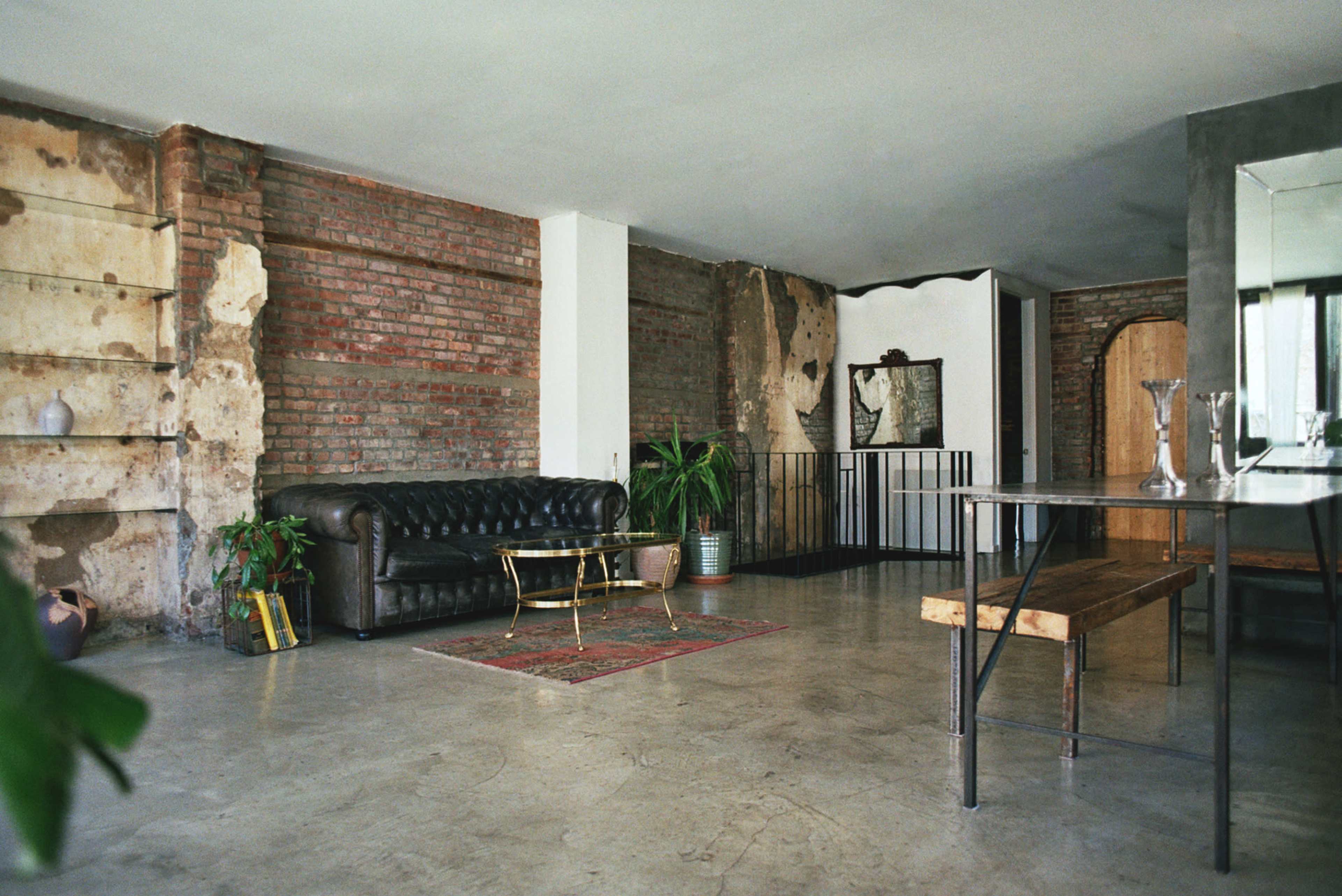 The image shows a spacious interior with exposed brick walls, a leather sofa, a wooden dining table, and various plants.