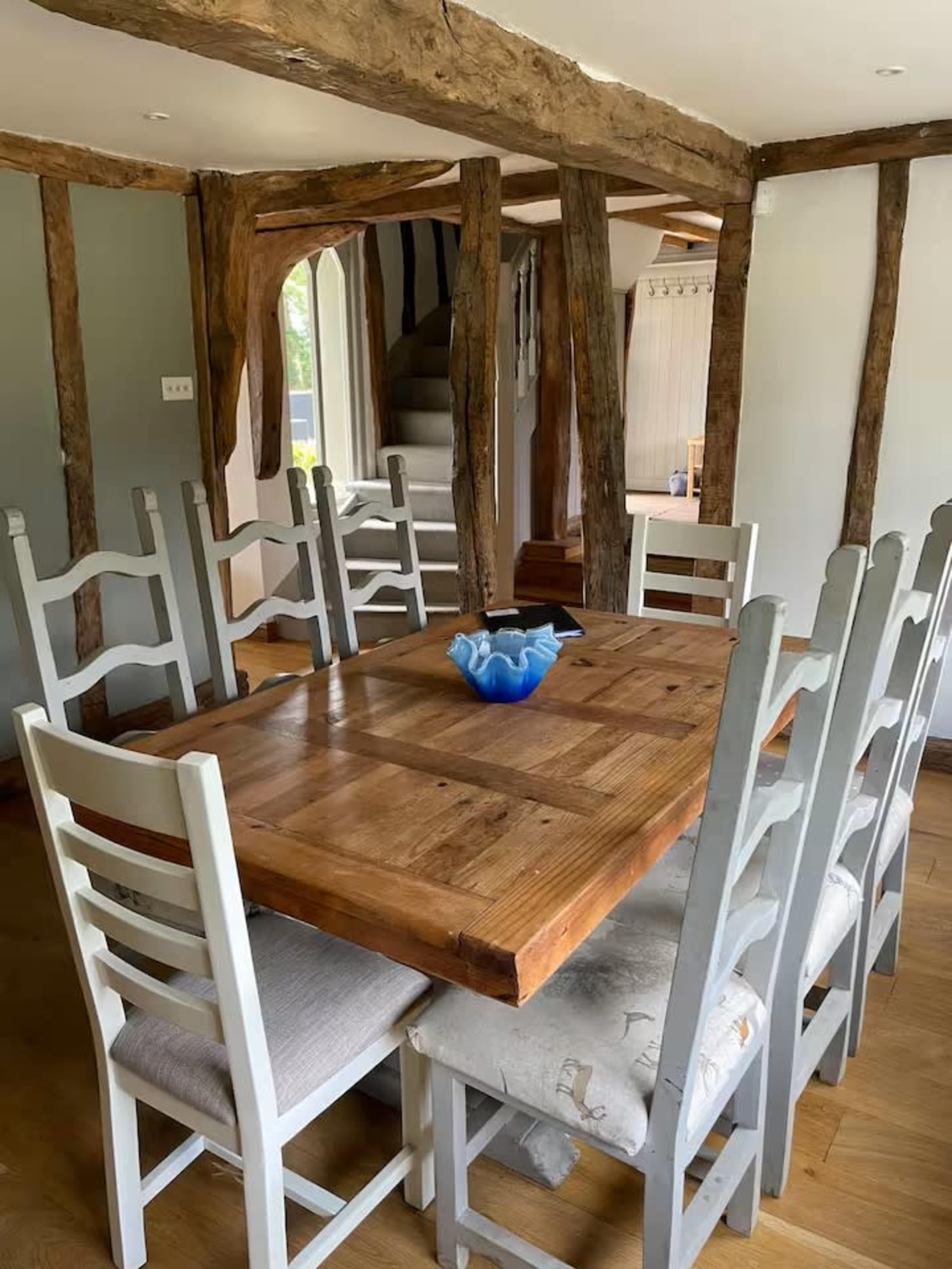 The image shows a dining area with a wooden table surrounded by white chairs, set within a room featuring exposed wooden beams and light-colored walls.