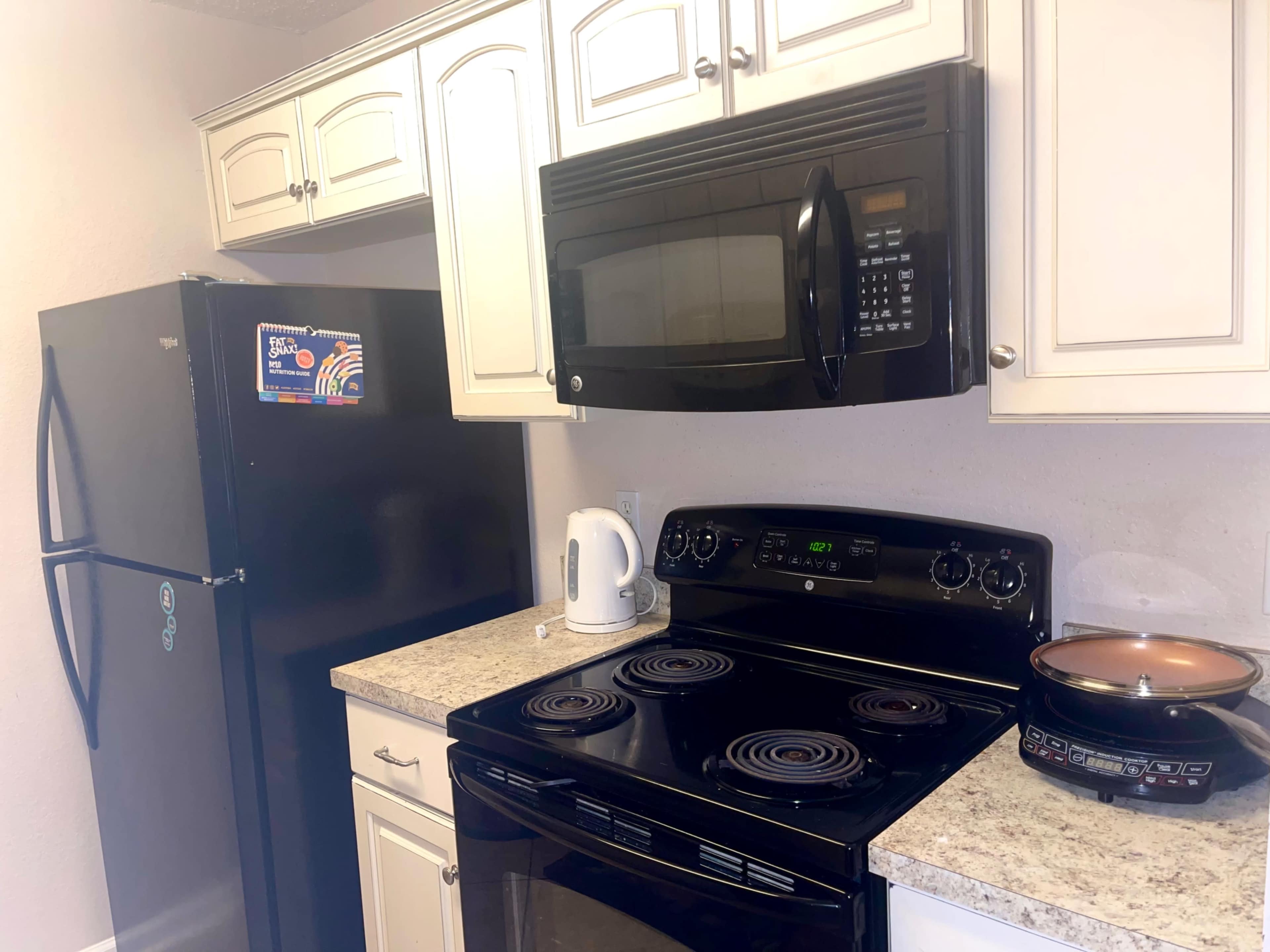 A kitchen with a black refrigerator, a microwave above a black stovetop, a kettle, and a multi-cooker on the countertop, all set against light-colored cabinets and a granite-like surface.