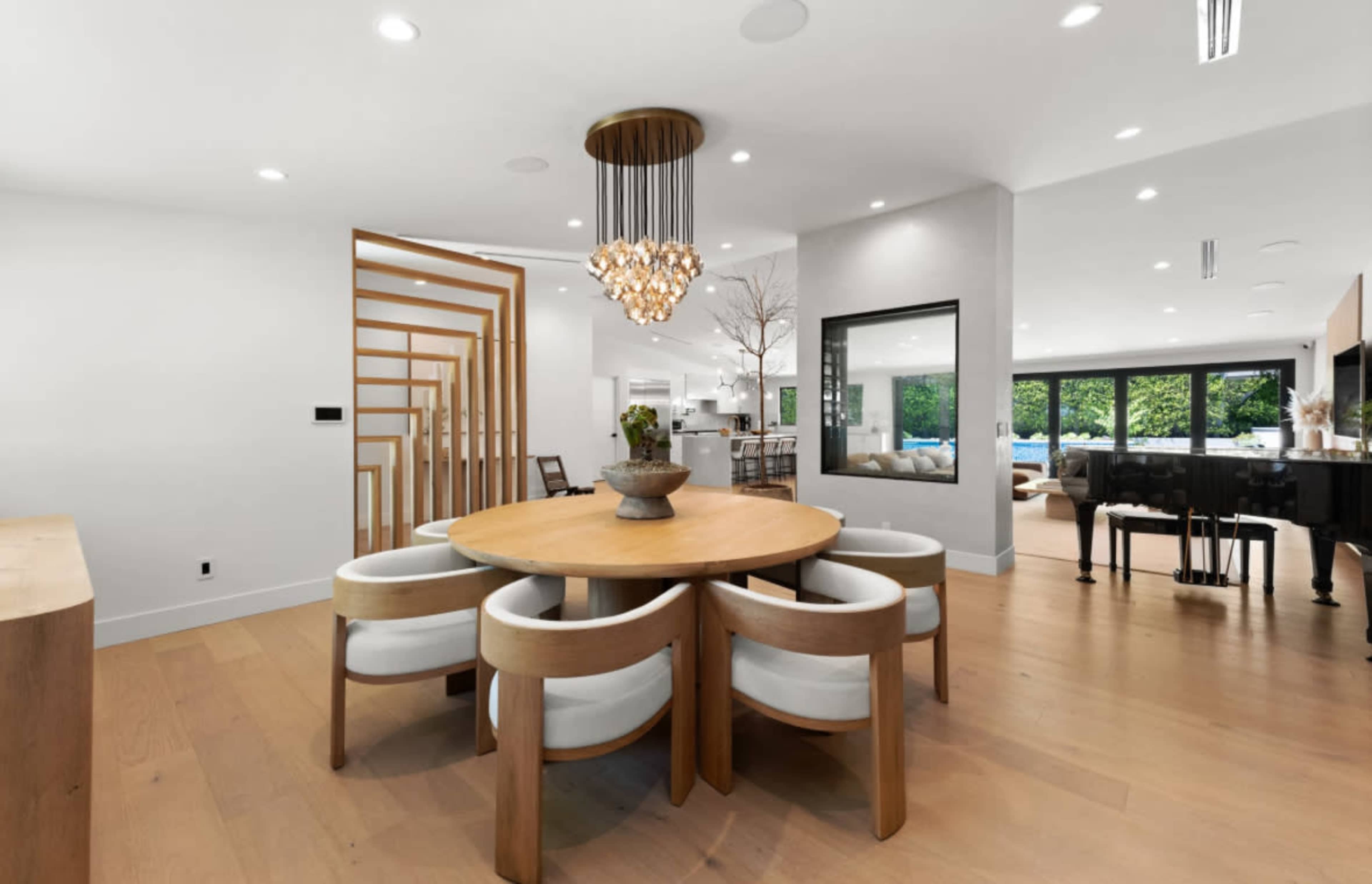 A round wooden dining table with curved chairs is set in a bright, open space featuring modern decor and a view of greenery through large windows.